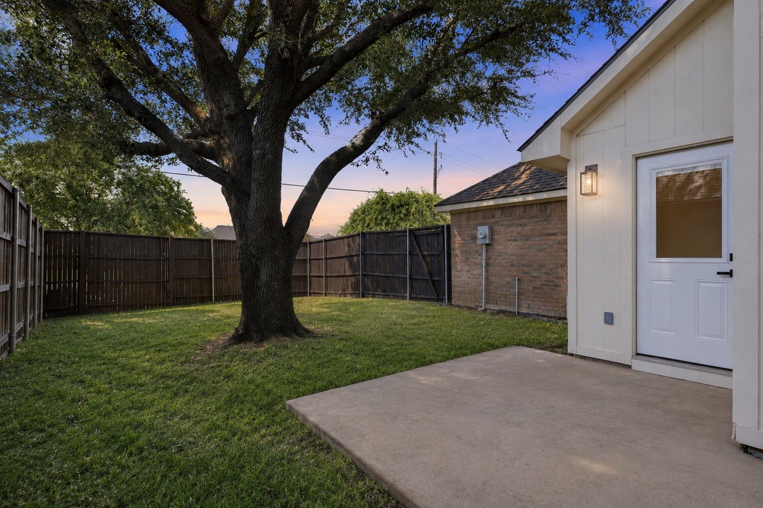 Fenced backyard with a large shade tree, lawn, and paved patio beside a white exterior door.