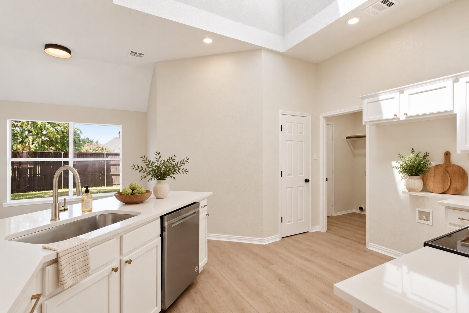 Bright white kitchen with a window above the sink, stainless-steel dishwasher, and light wood-look floors.