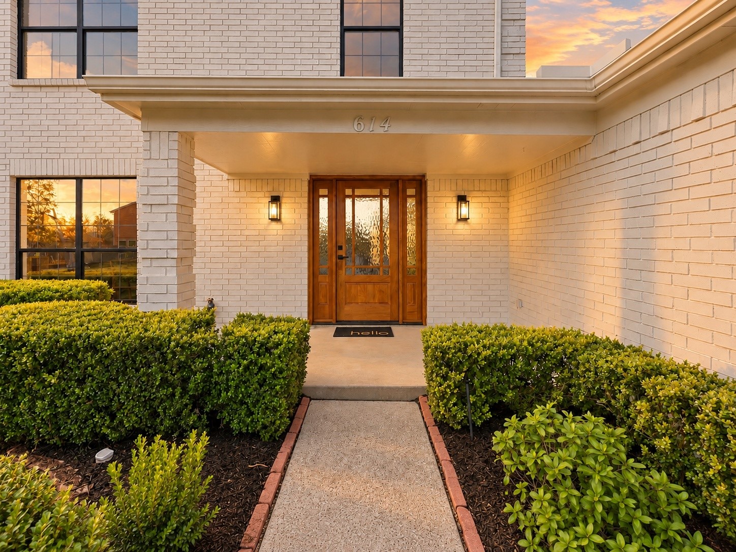 White brick home entrance with a covered porch, wood-and-glass double door, and neatly trimmed hedges.
