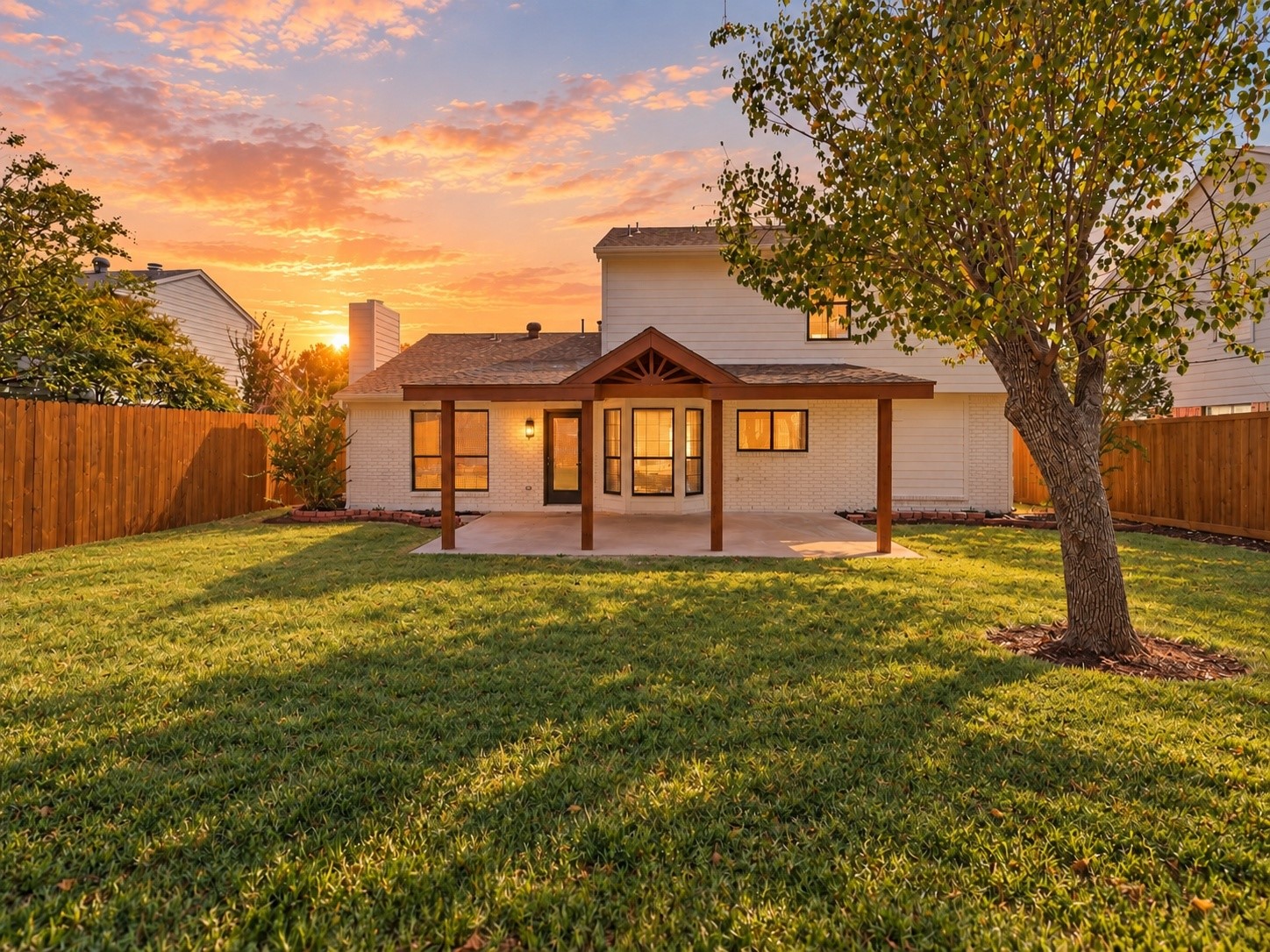Rear of a two-story white house with a covered patio and fenced lawn at sunset.