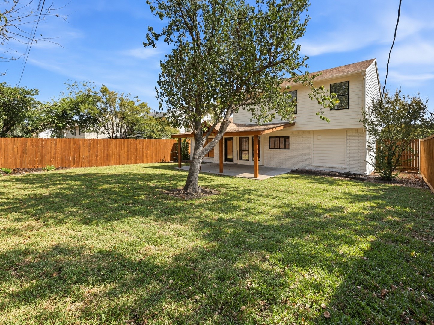 Two-story white house and covered patio behind a broad grassy yard and wooden privacy fence.
