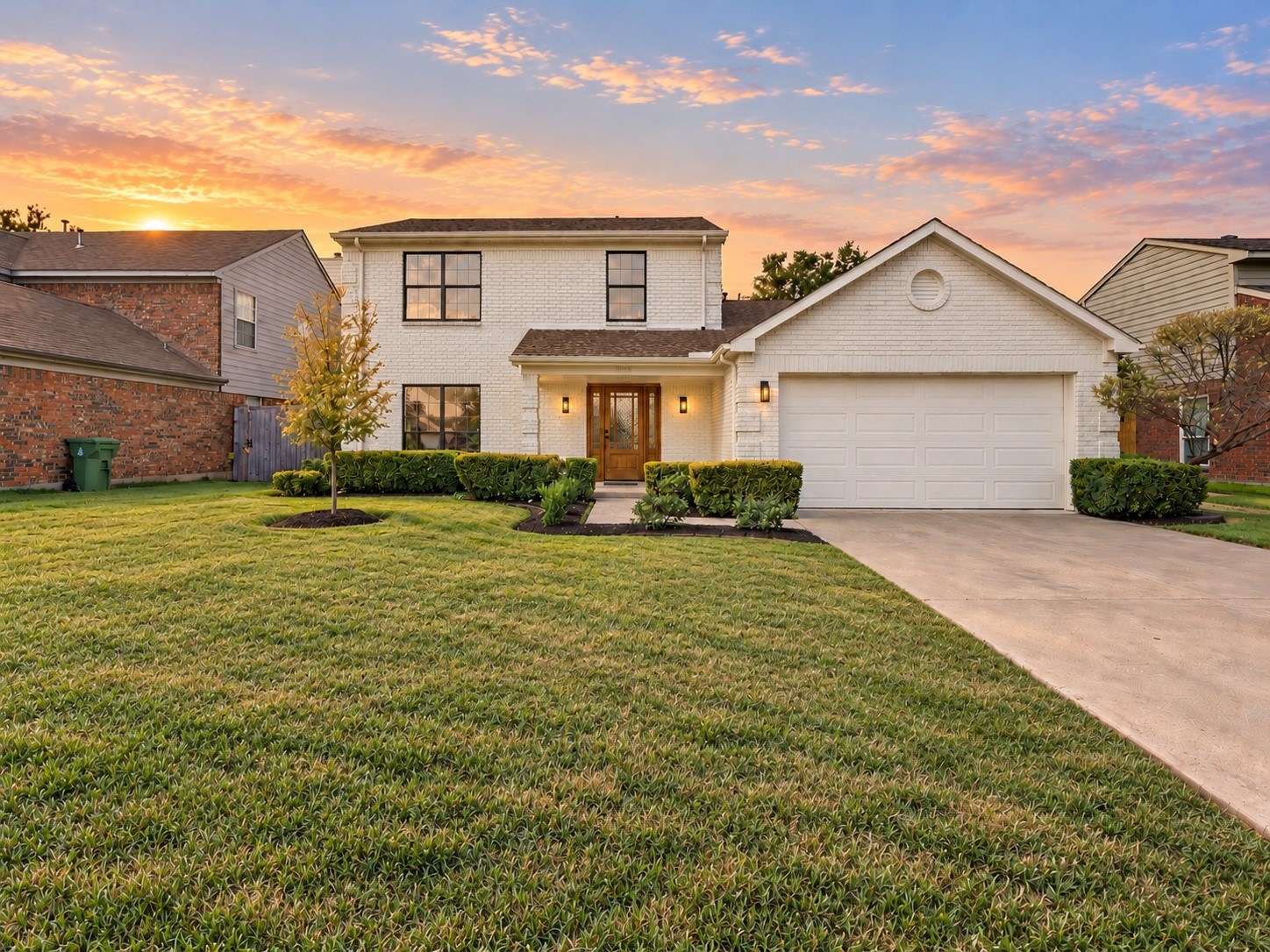 Two-story white brick home with an attached garage, landscaped lawn, and colorful sunset sky.
