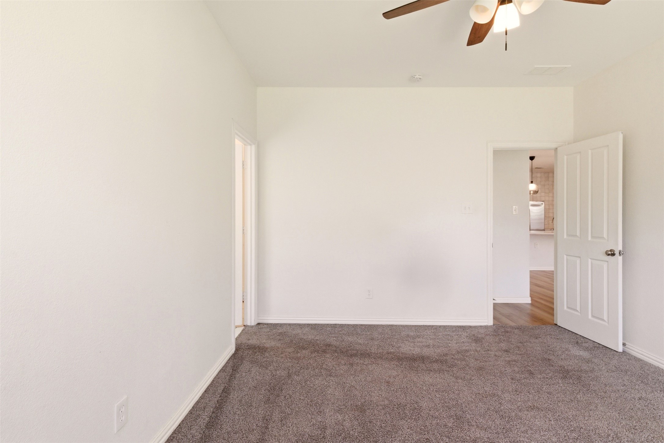 Empty bedroom with gray-brown carpet, a ceiling fan, and an open doorway to a wood-floored area.