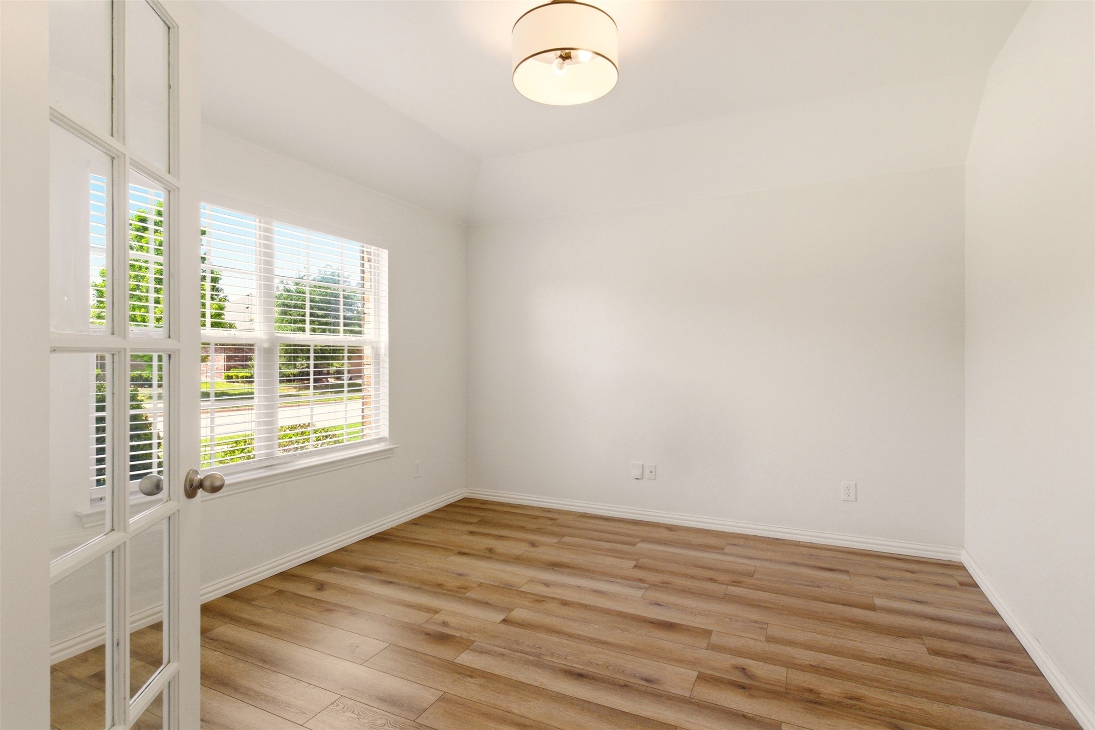 Vacant room with wood-look flooring, a large window with blinds, and a glass-paneled door.