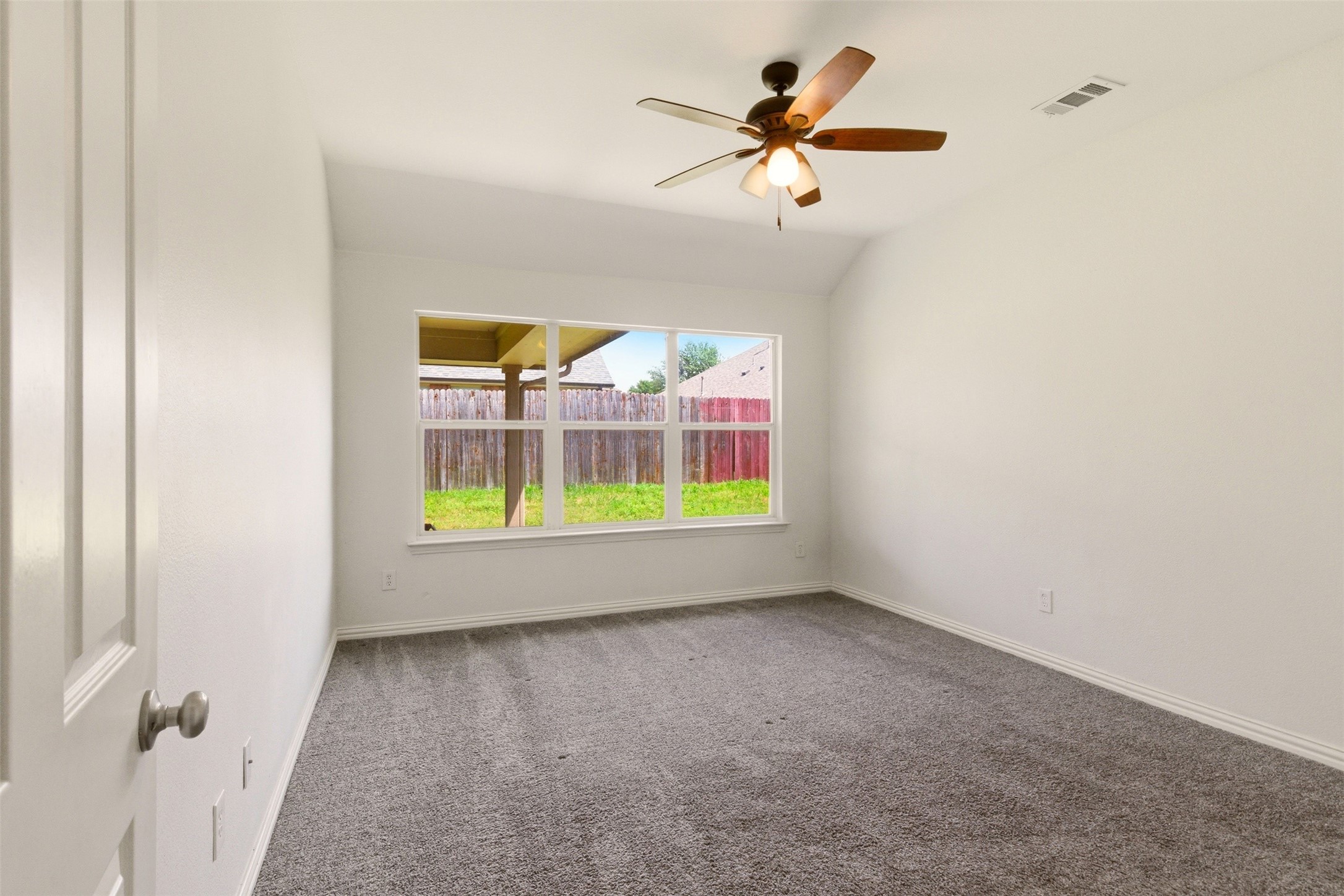 Vacant bedroom with gray carpet, a ceiling fan, and a large window overlooking a fenced yard.