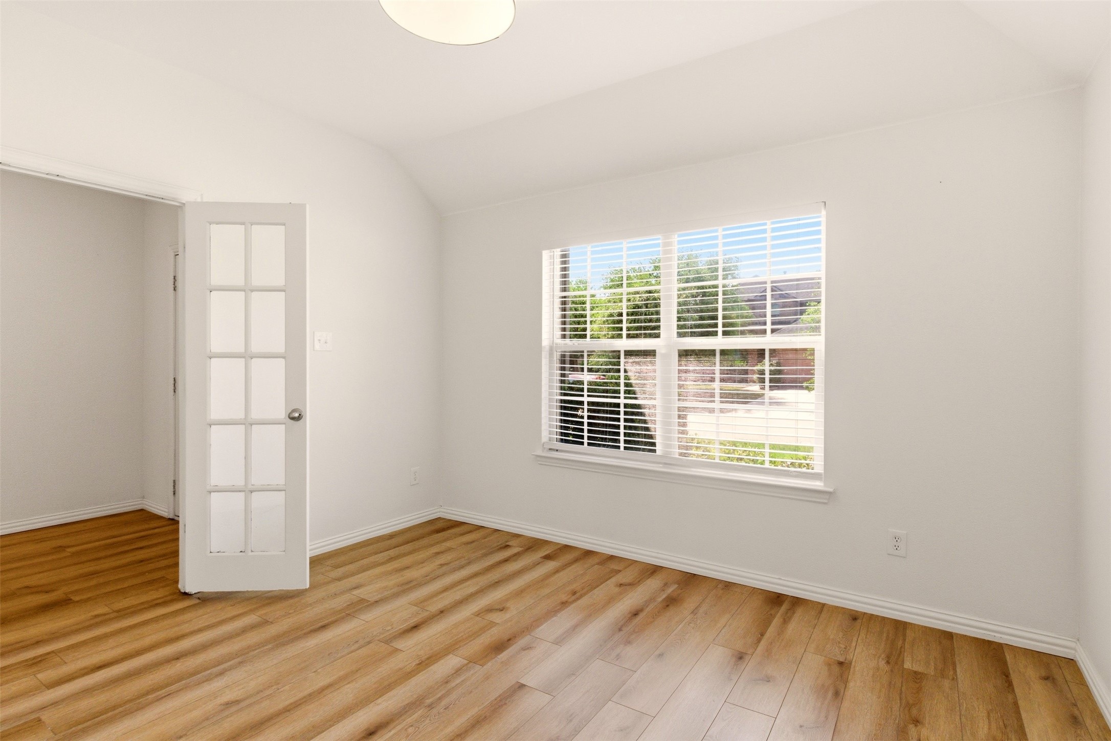 Empty room with light wood-look flooring, a multi-pane door, and a large window with blinds.