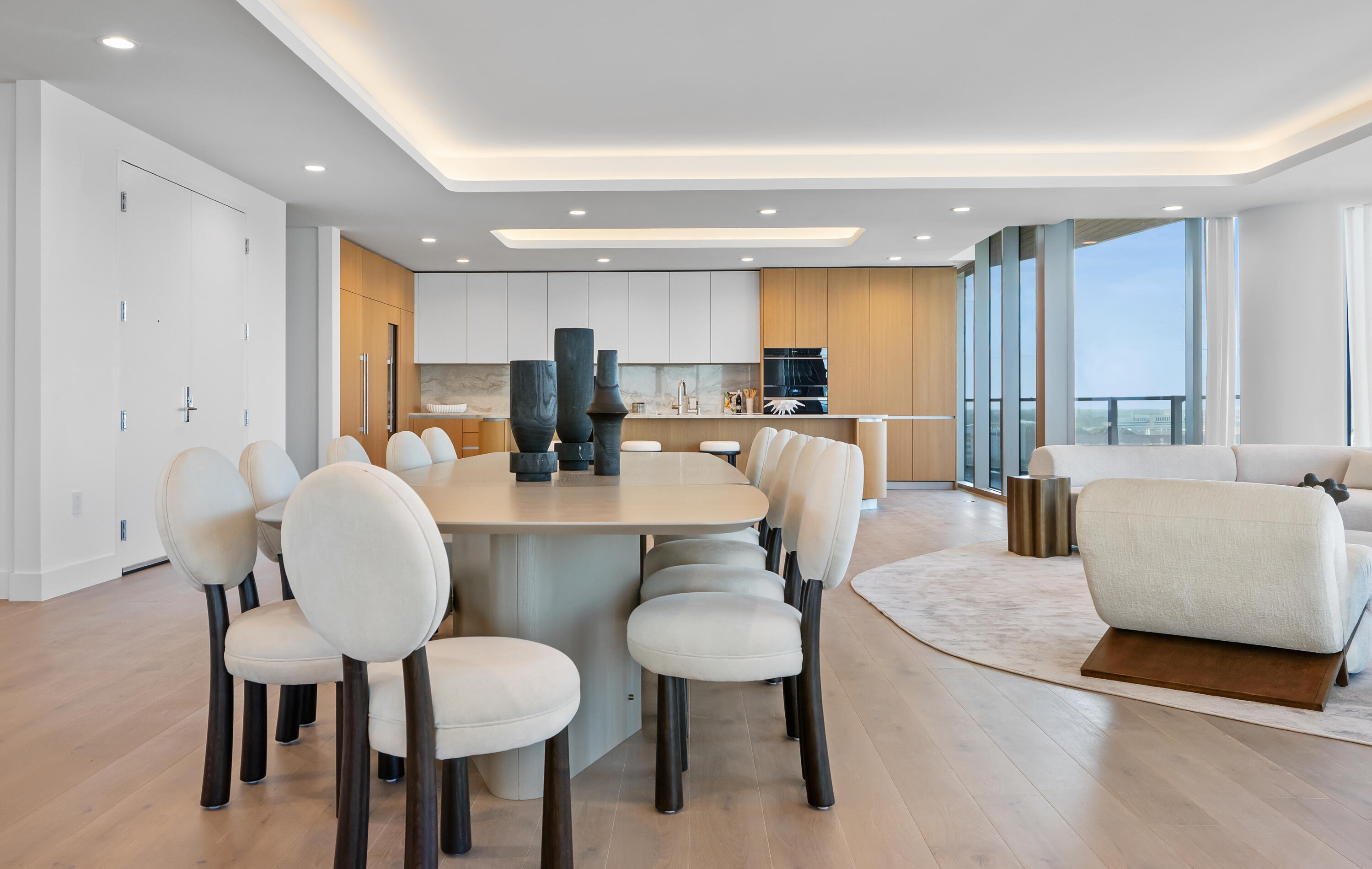 Open-plan dining area with upholstered chairs, a light wood kitchen, and floor-to-ceiling windows.