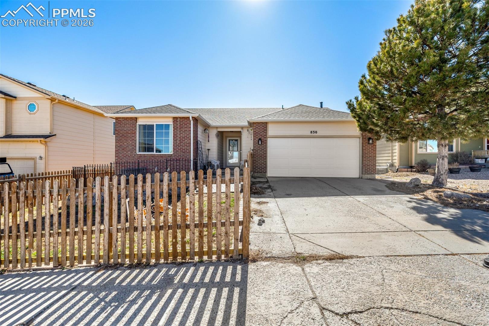 Single-story brick-and-siding home with an attached garage, picket fence, and large pine tree.