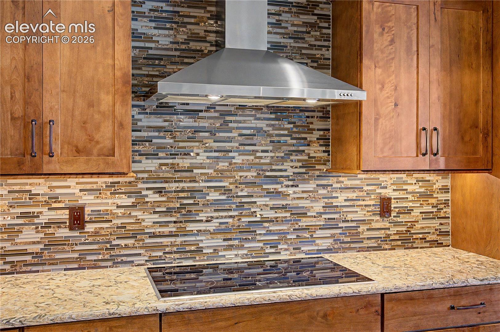 Kitchen with stainless steel range hood, multicolored mosaic backsplash, wood cabinets, and stone counters.