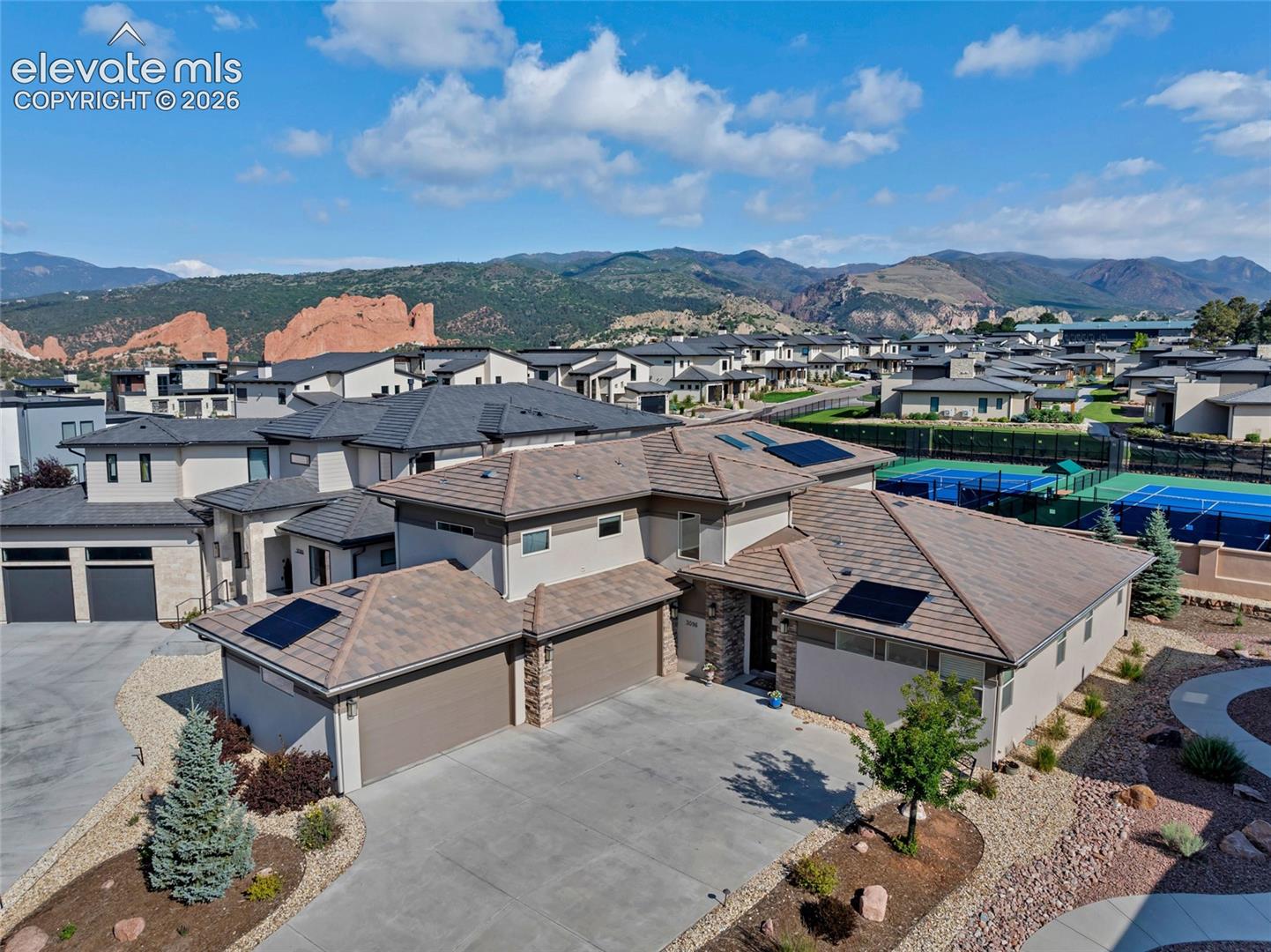 Aerial view of a modern home with rooftop solar panels, tennis courts, and red rock formations against mountain ridges.