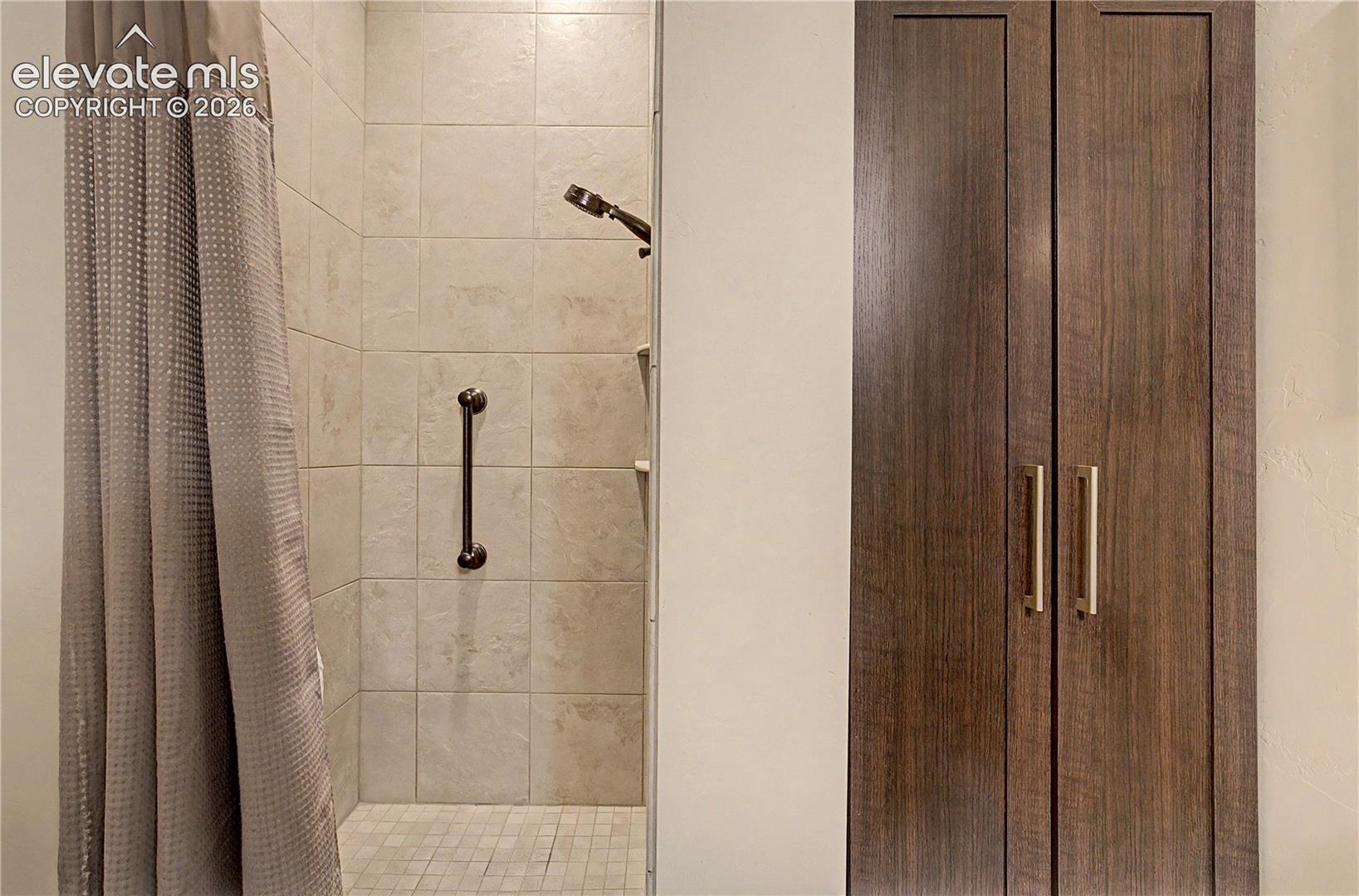 Bathroom shower with beige tile and grab bar beside a tall dark wood cabinet.