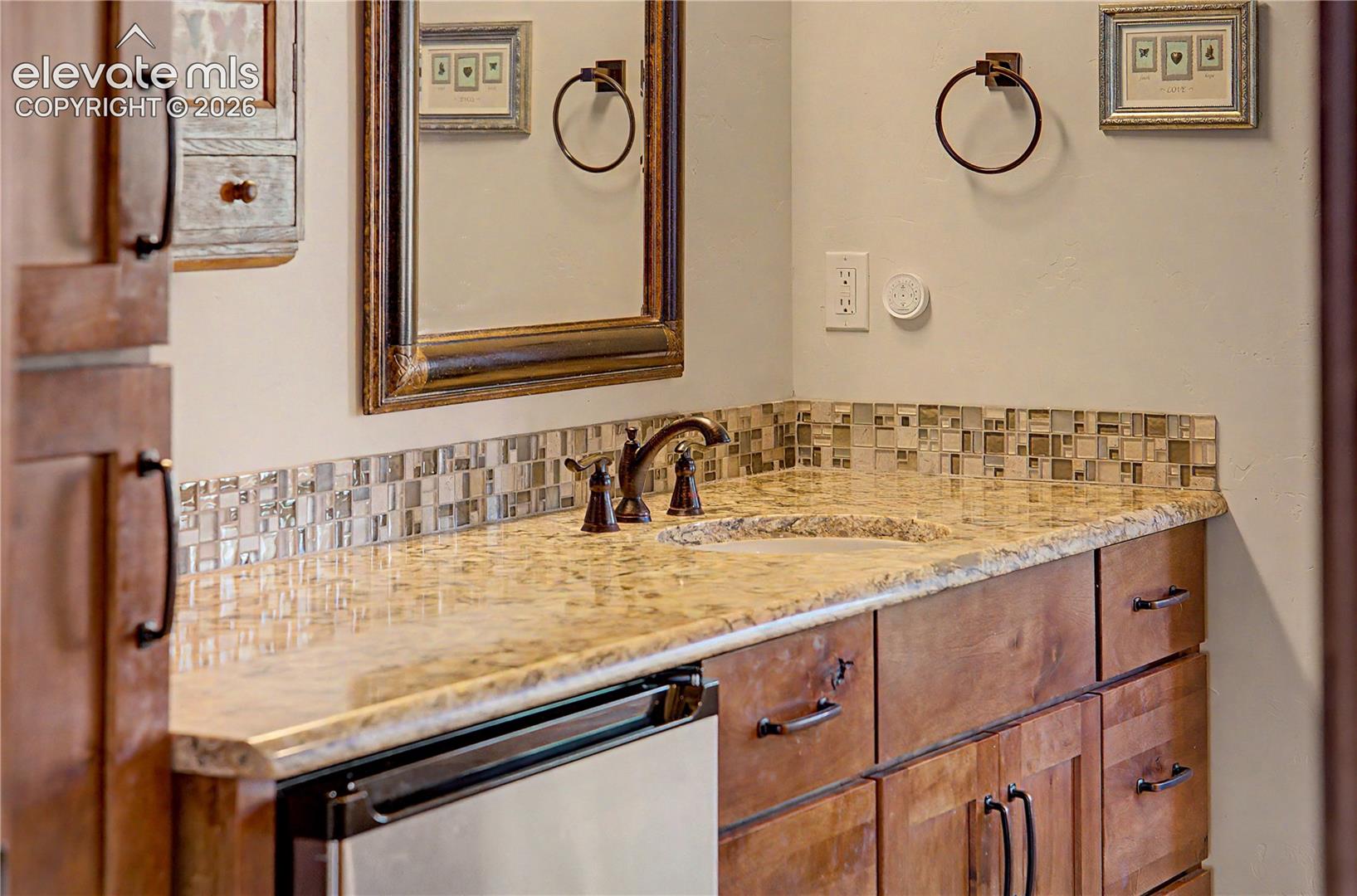 Bathroom vanity with a stone countertop, mosaic backsplash, dark bronze faucet, and wood cabinetry.