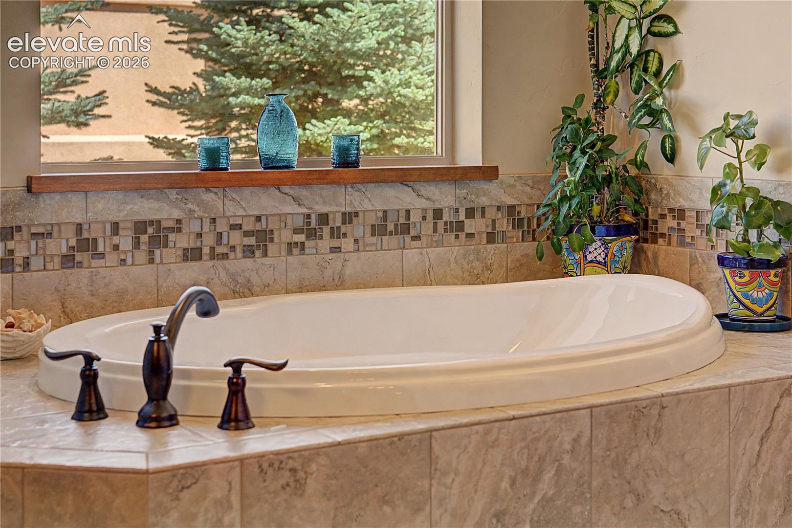 Oval soaking tub with stone tile surround, mosaic backsplash, dark faucet, and plants beside a window.