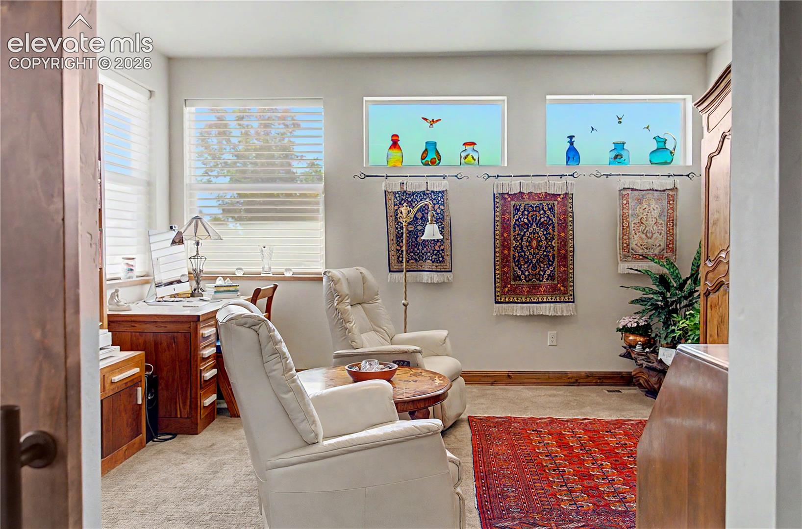 Furnished home office with cream recliners, a wooden desk, colorful glass ornaments, and patterned rugs.