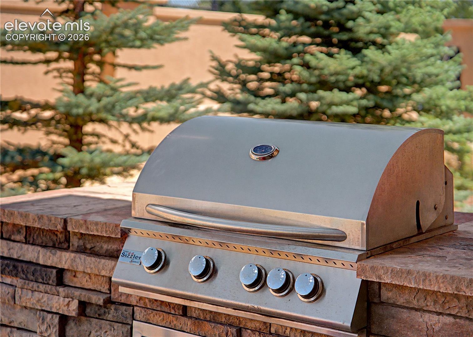 Stainless steel gas grill set into a stacked-stone outdoor counter beneath evergreen trees.