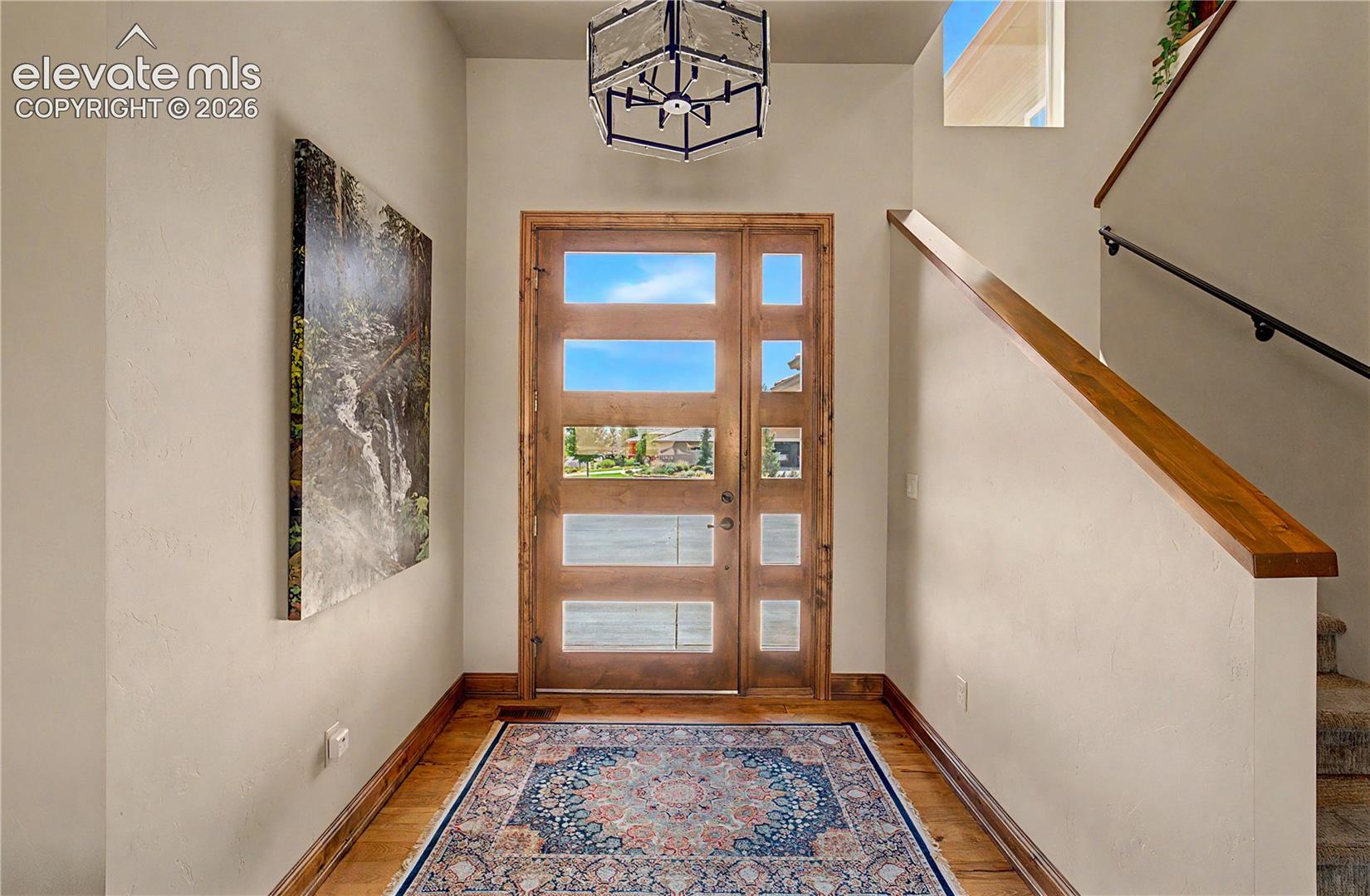 Entry foyer with a wood-and-glass front door, patterned rug, and staircase with wood handrails.