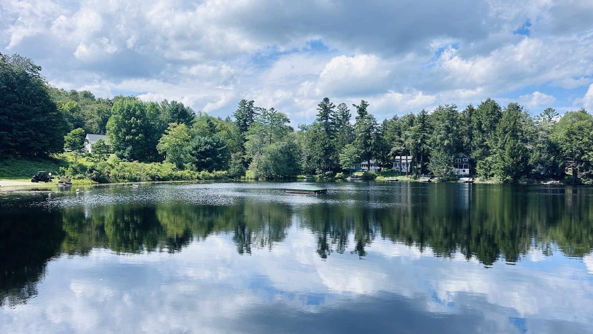 Lake with wooded shoreline homes and a floating dock reflected beneath a cloudy blue sky.