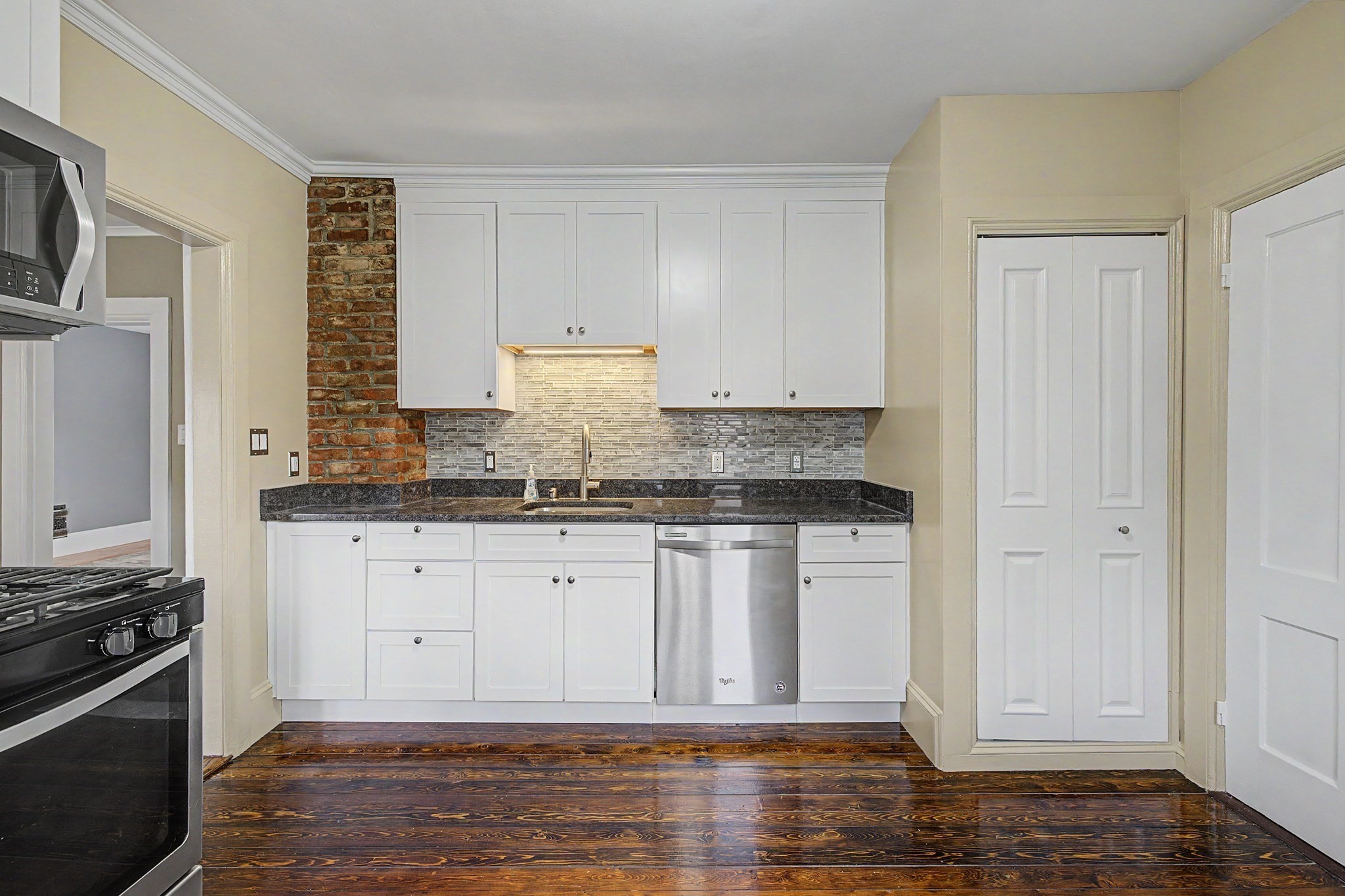 Kitchen with white cabinetry, stainless steel appliances, exposed brick, and dark polished wood floors.