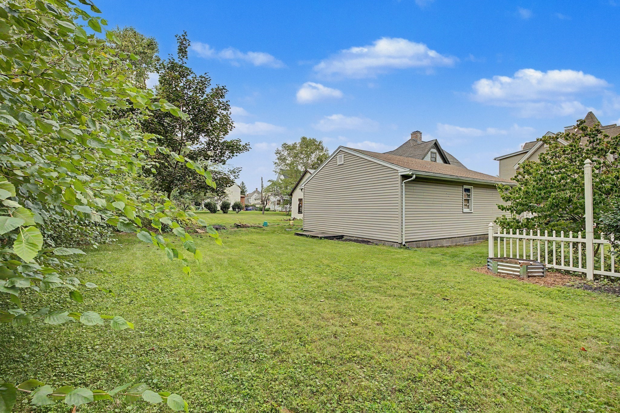 Beige-sided house beside a broad grassy yard, leafy trees, and a white picket fence.