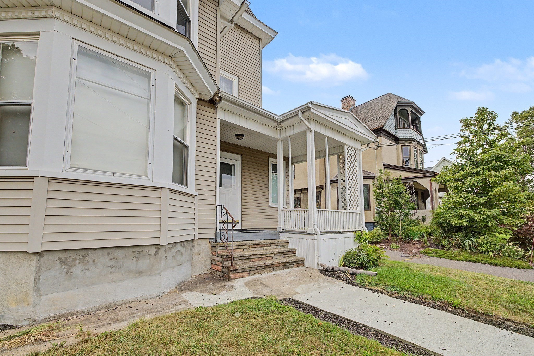 Beige-sided home with a white covered porch, lattice panels, and stone steps beside a concrete walkway.