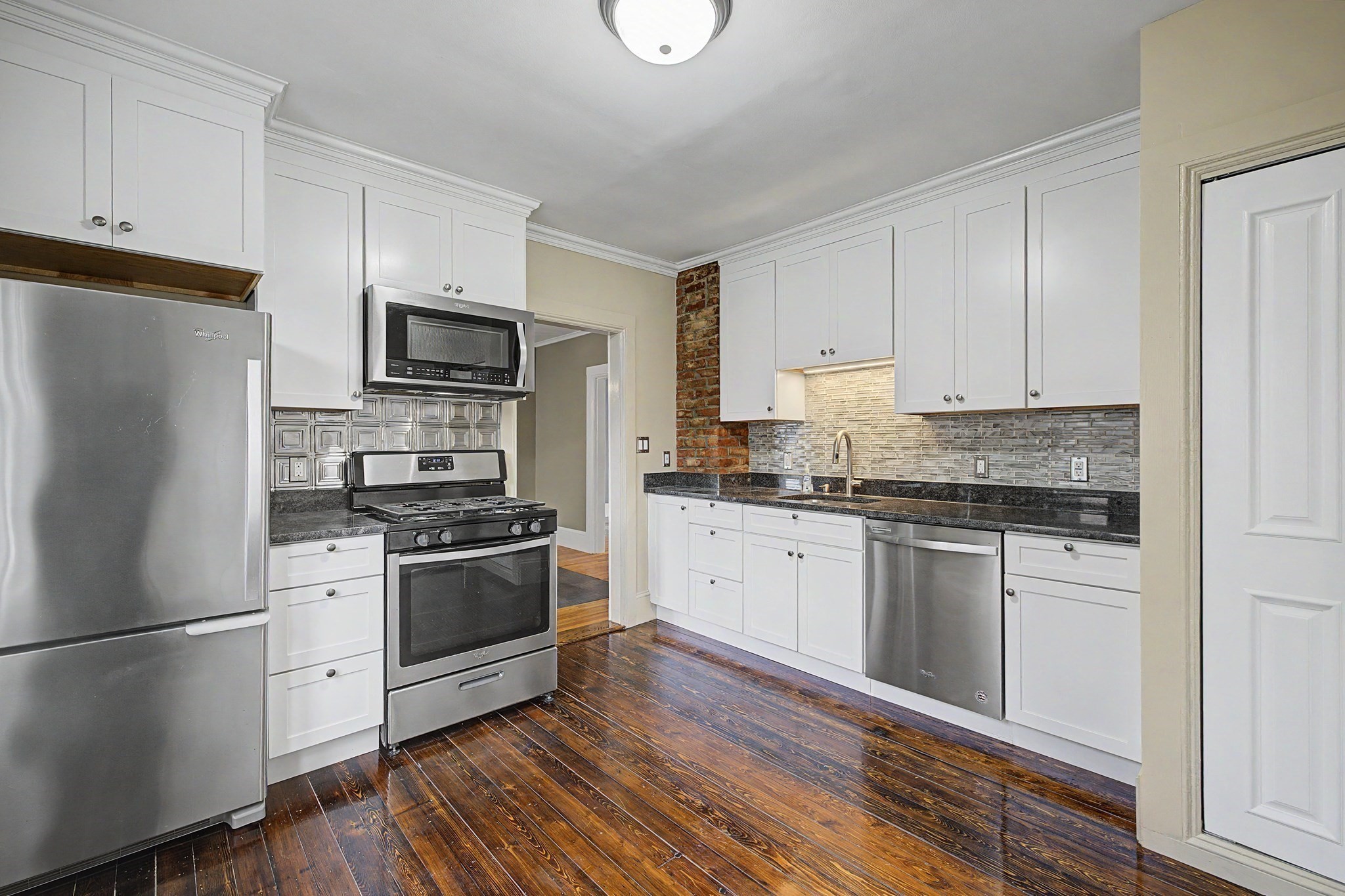 Kitchen with white cabinets, stainless steel appliances, mosaic backsplash, exposed brick, and dark hardwood floors.