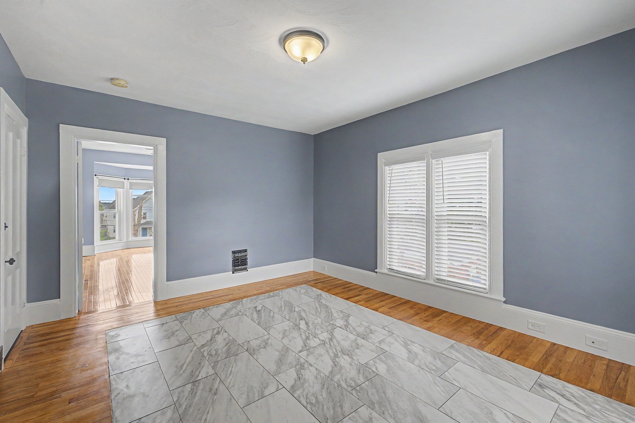 Empty gray room with marble-pattern tile bordered by hardwood and shuttered windows.