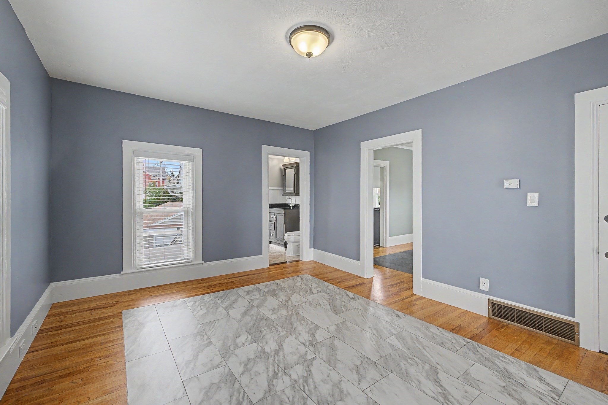 Empty blue-gray room with hardwood floors, a marble-look tile section, and doorways to a bathroom and hall.