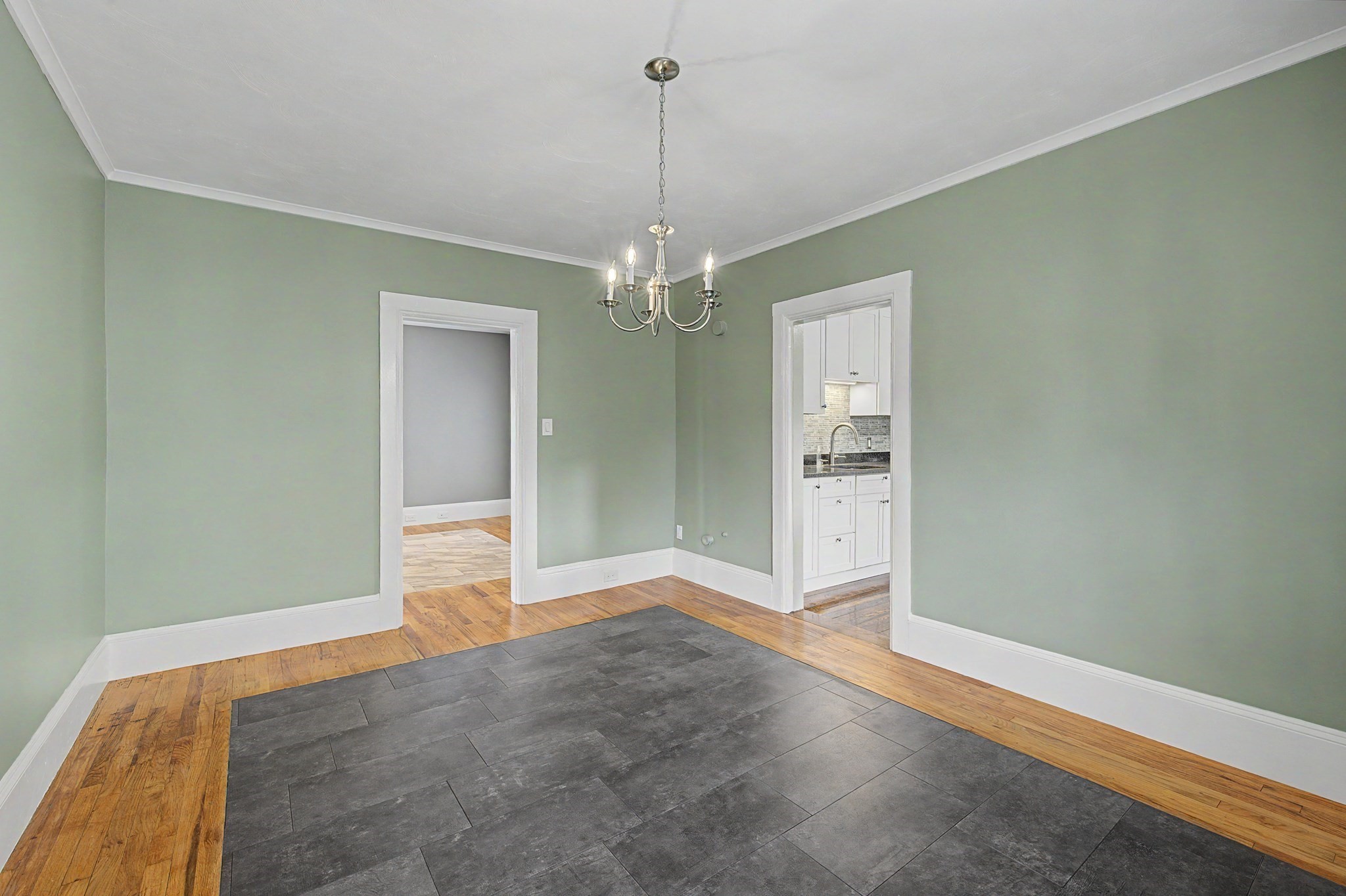 Empty sage-green room with a chandelier, gray tile inset in hardwood, and a doorway to the kitchen.