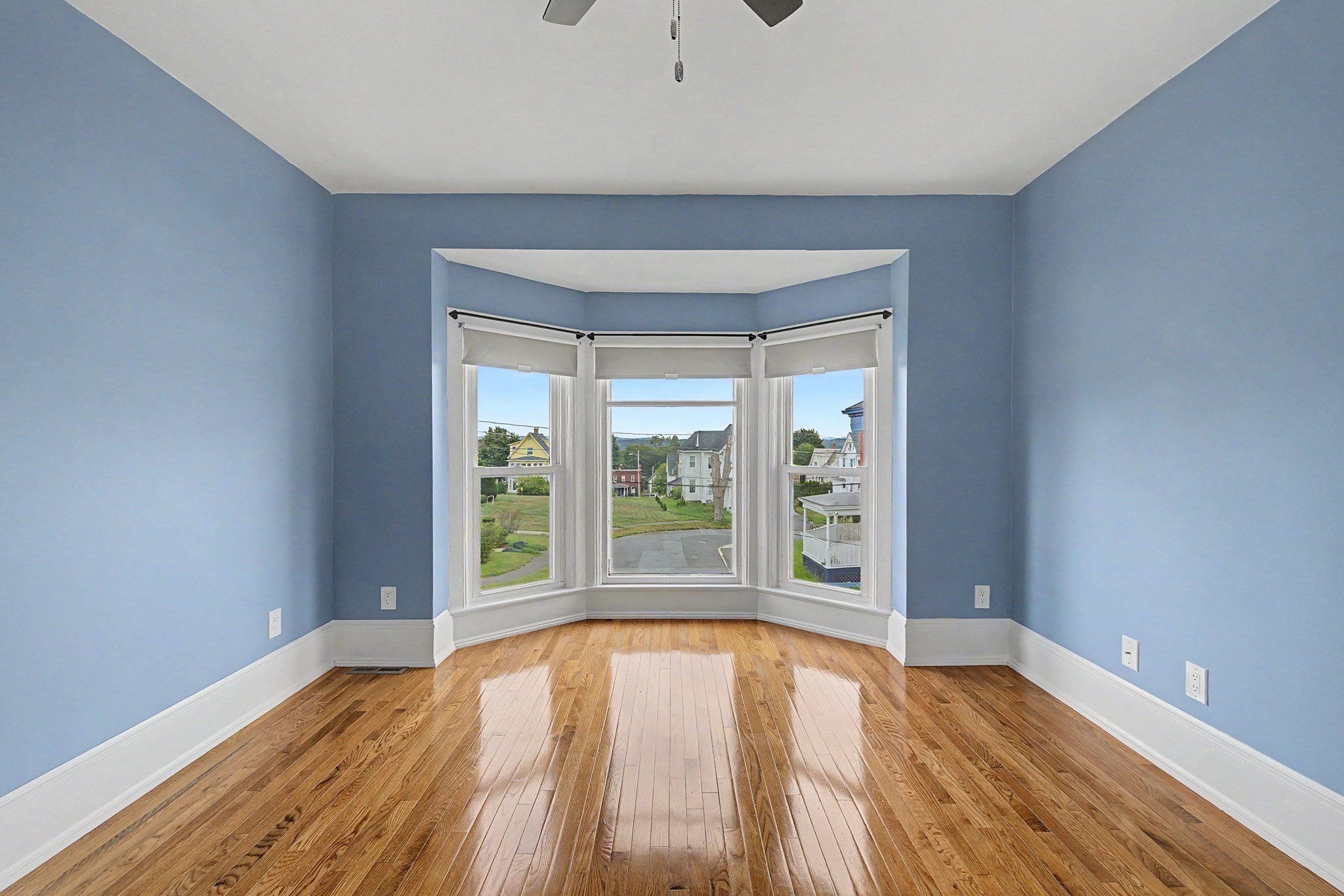 Empty blue room with polished hardwood floors and a bay window overlooking the neighborhood.
