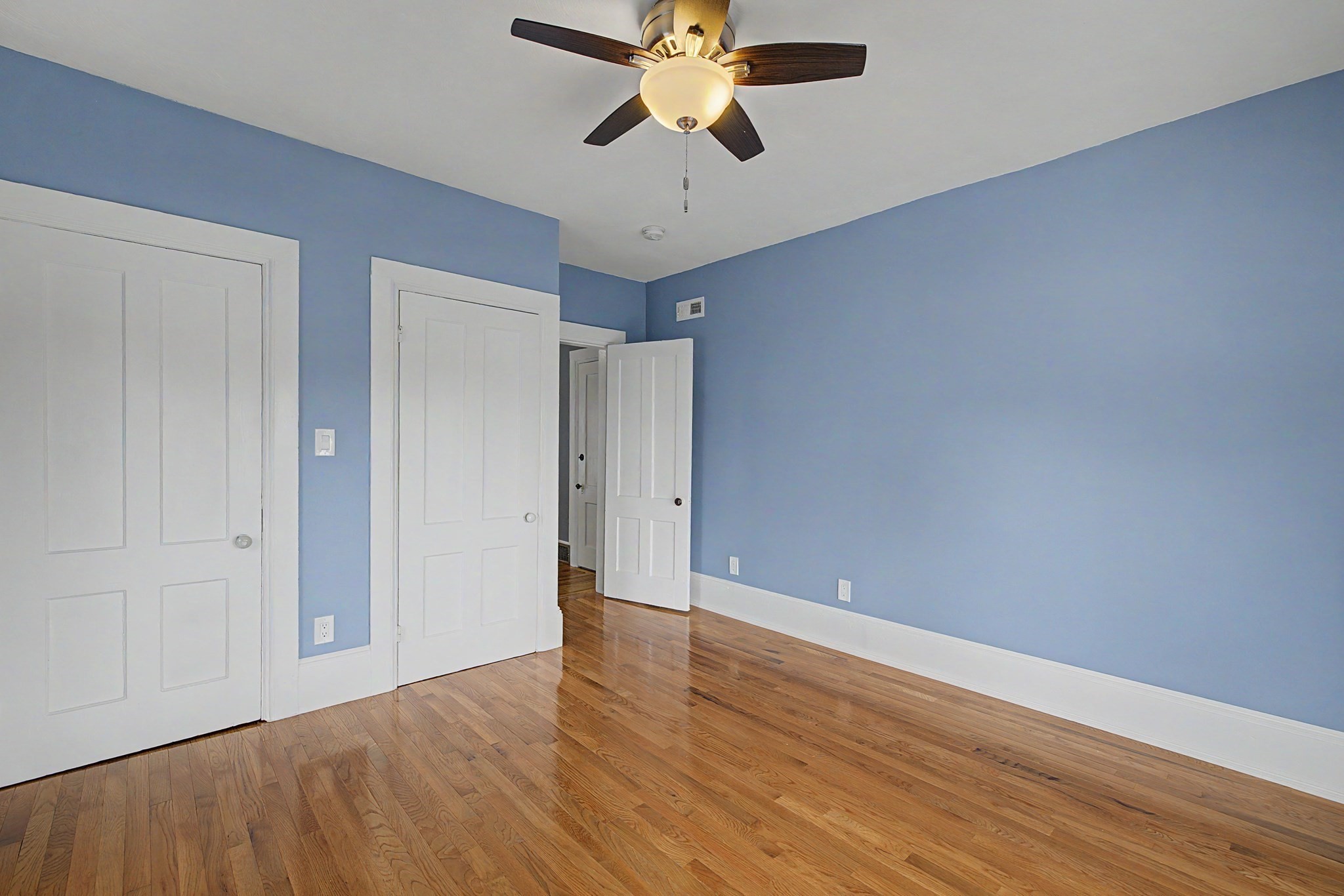 Empty bedroom with blue walls, hardwood floors, white paneled doors, and a ceiling fan.