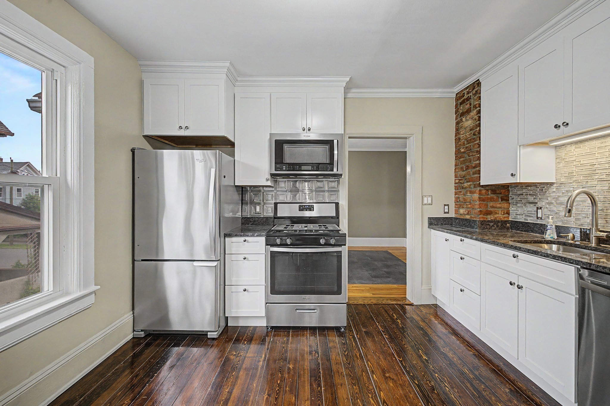 Kitchen with white cabinetry, stainless steel appliances, exposed brick, and dark hardwood floors.