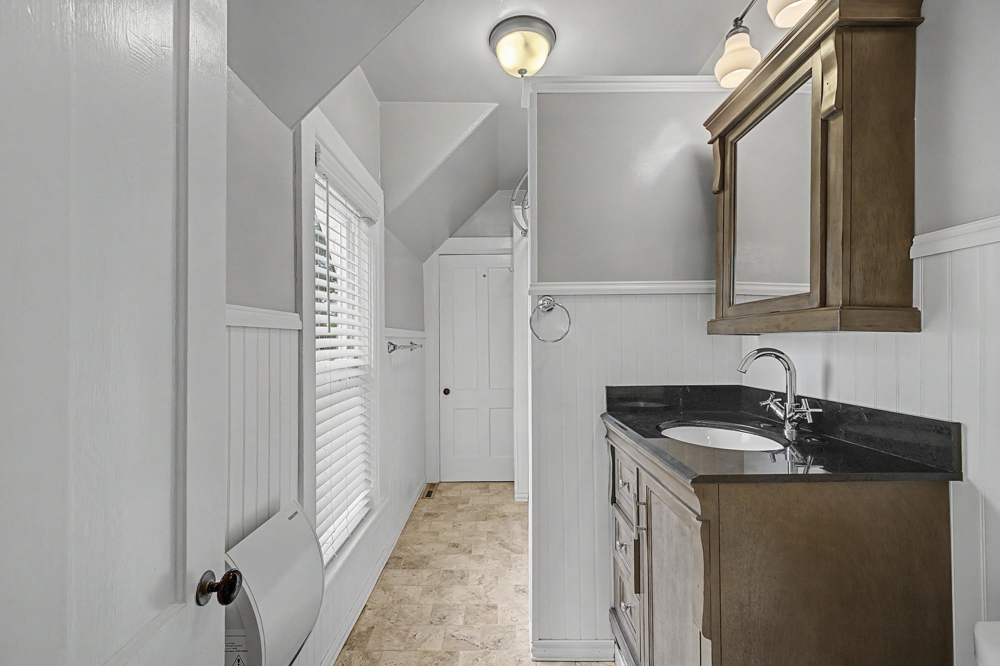 Narrow bathroom with a dark countertop, wood vanity, mirrored cabinet, and window with blinds.