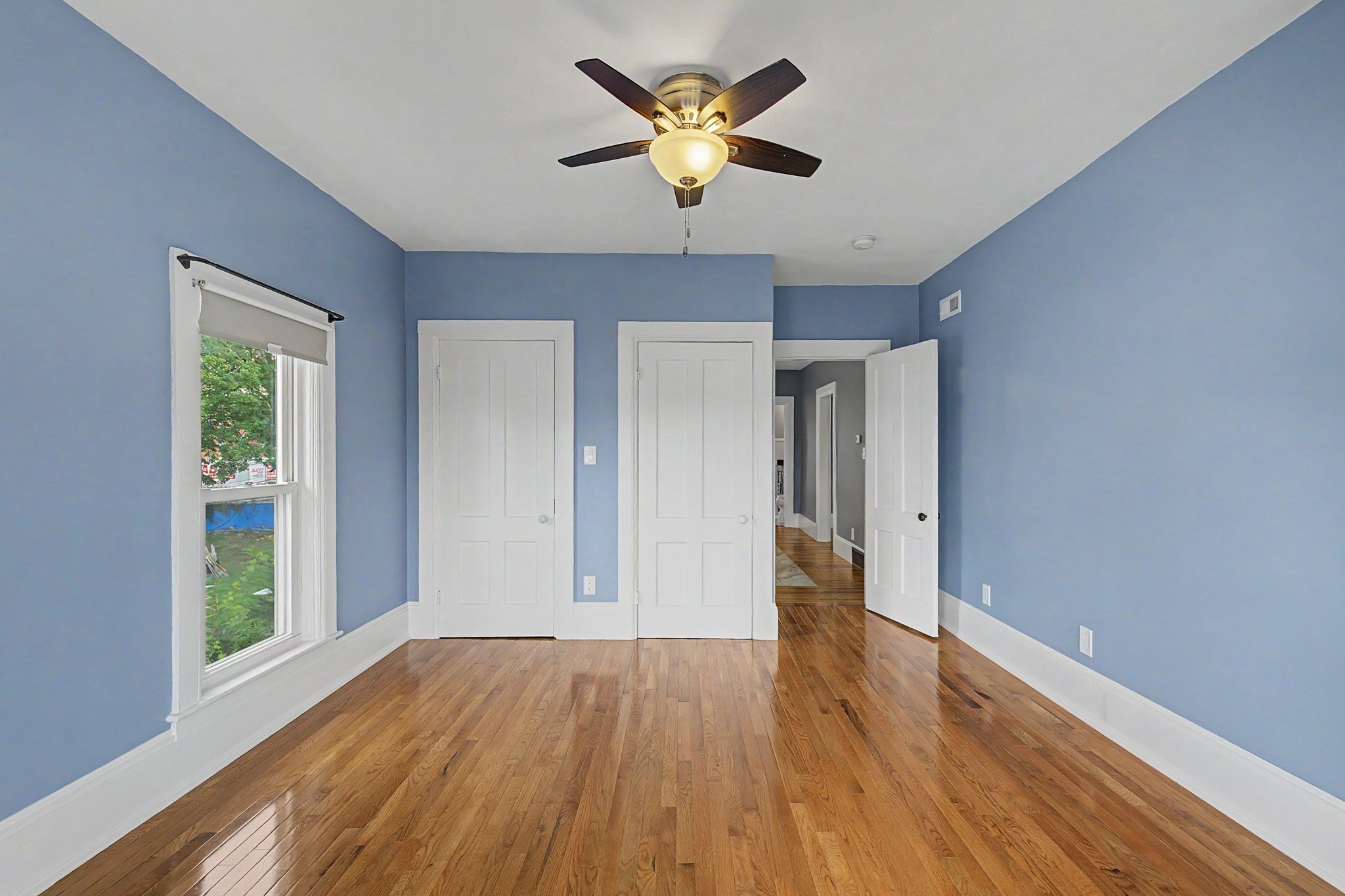 Empty blue bedroom with hardwood floors, two closets, a large window, and a ceiling fan.