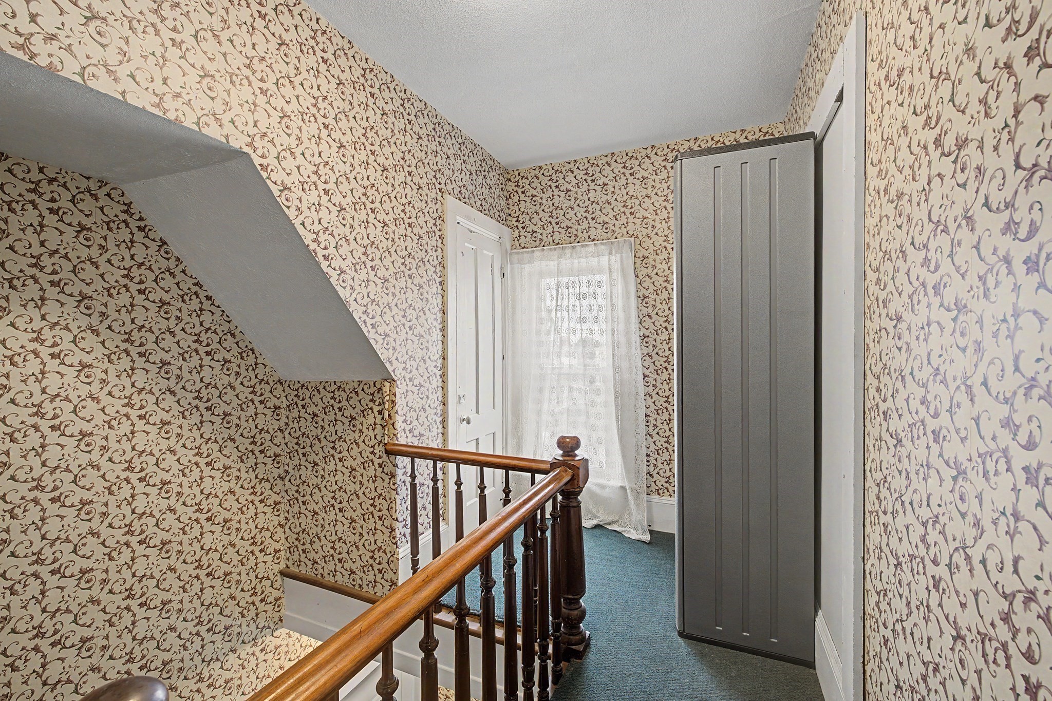 Stair landing with patterned wallpaper, blue carpet, a wooden banister, and a tall gray cabinet.