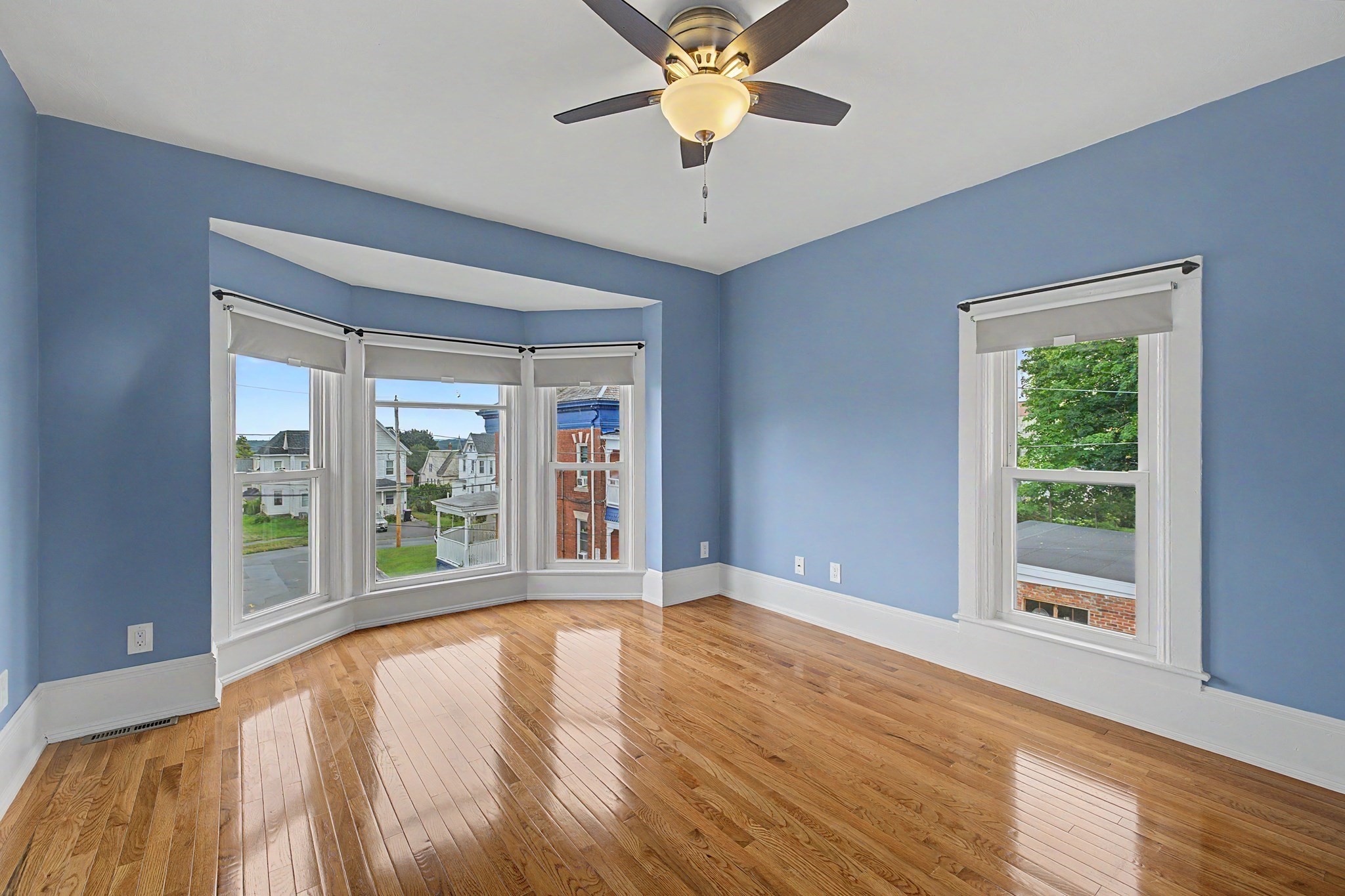 Empty blue-walled room with polished hardwood floors, bay windows, and a ceiling fan.