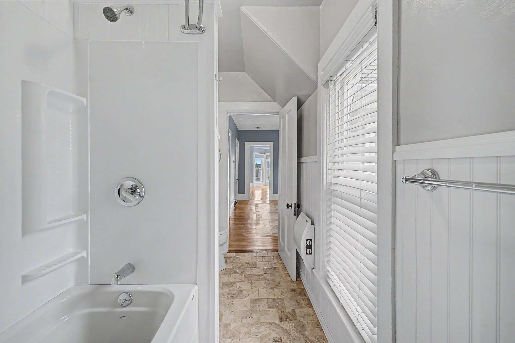 Narrow bathroom with a white tub and shower, tiled floor, and a hallway beyond.