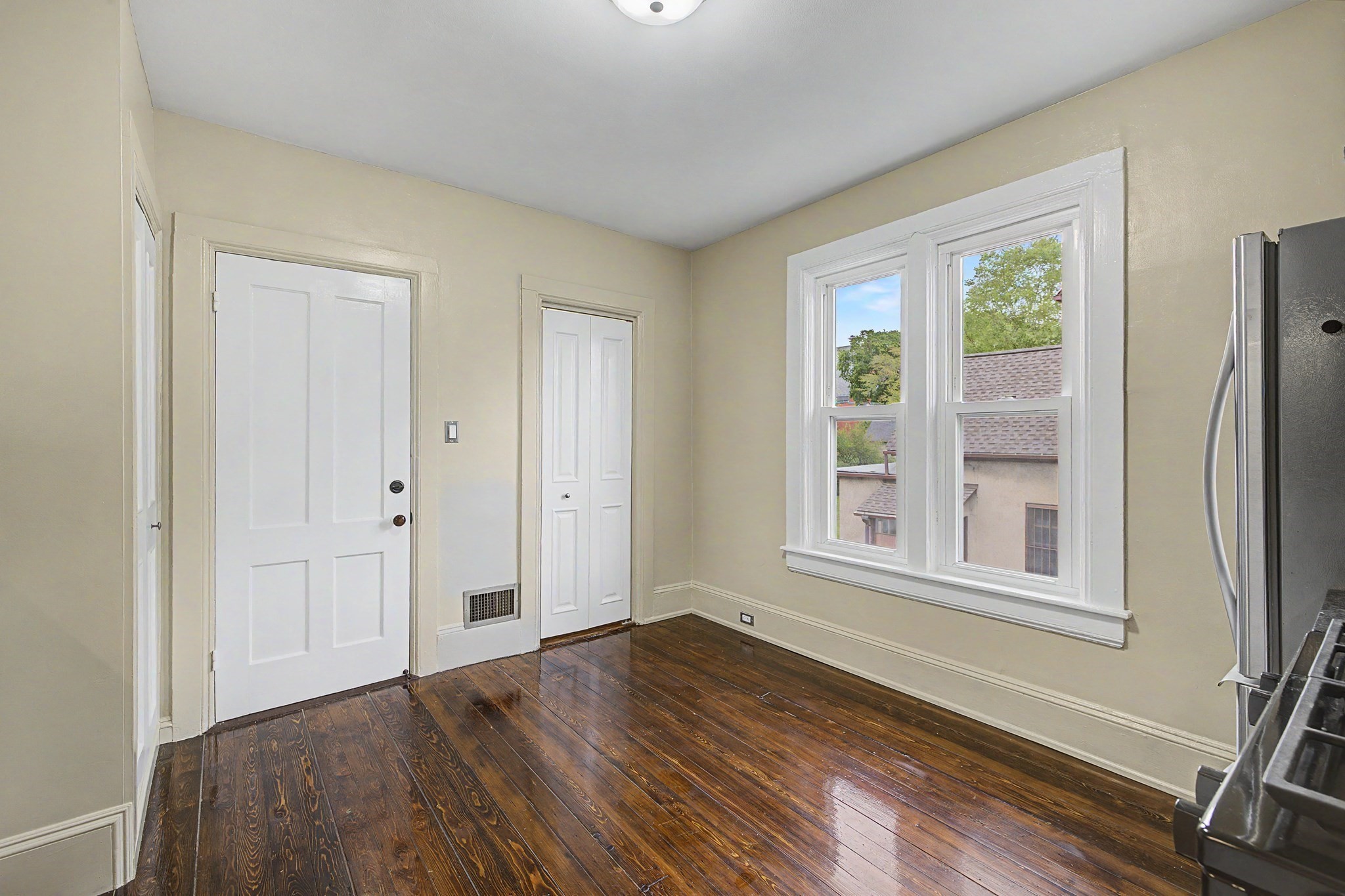 Vacant room with dark hardwood floors, white doors, a double window, and a stainless-steel refrigerator.