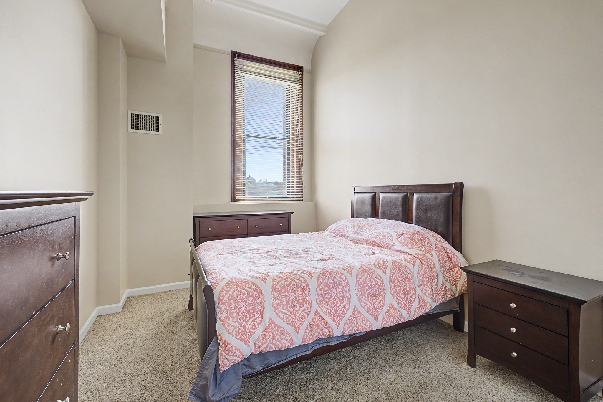 Bedroom with a dark wood bed, patterned pink bedding, and a tall window with blinds.