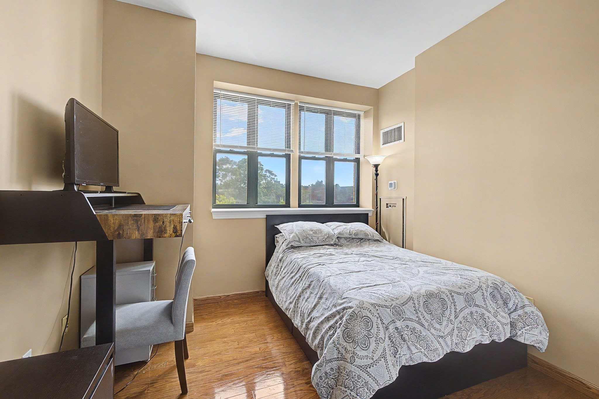 Bedroom with a patterned bedspread, tall windows, and a desk beside the bed.