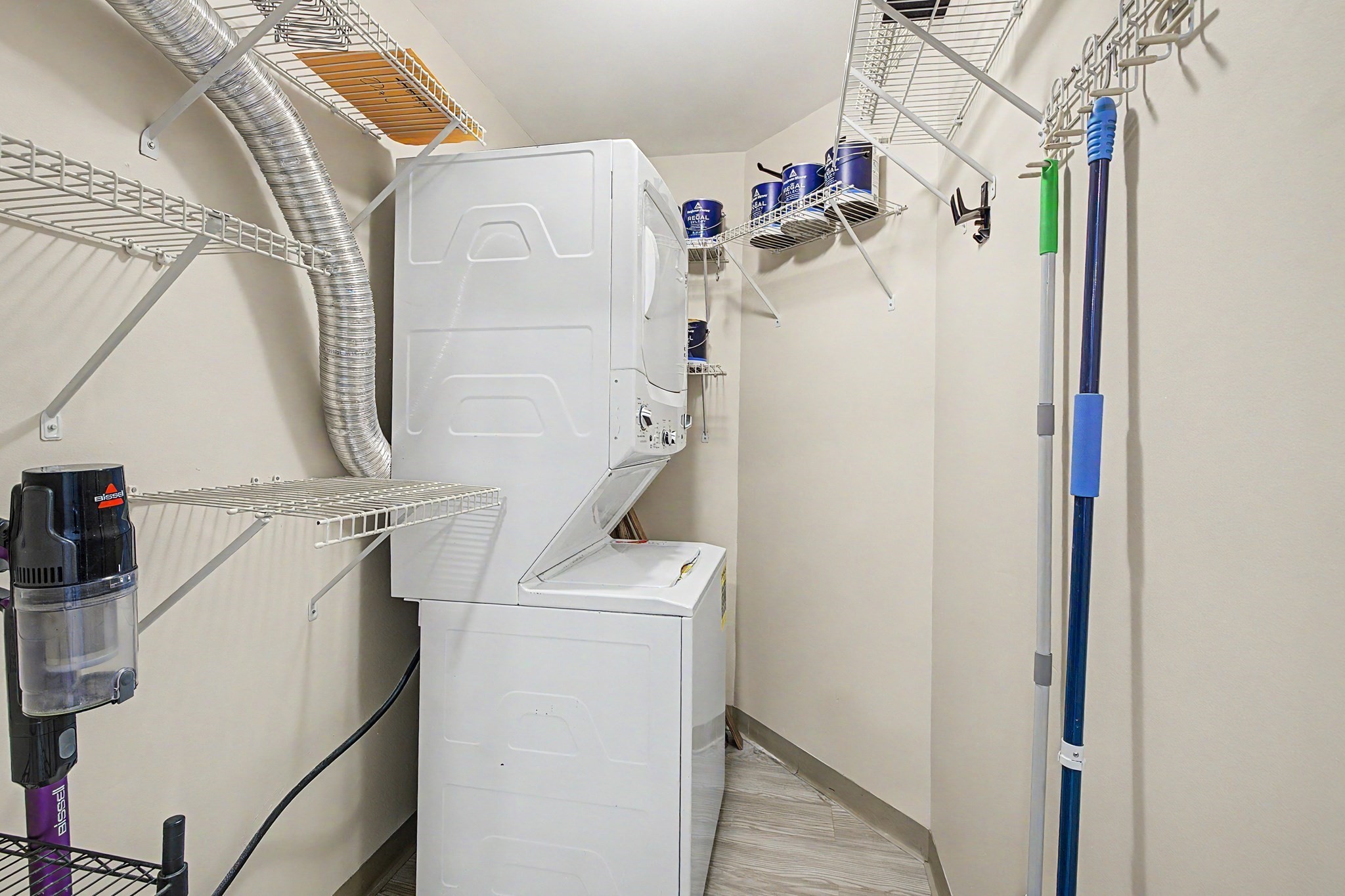 Laundry room with stacked washer and dryer, wire shelves, and cleaning tools.