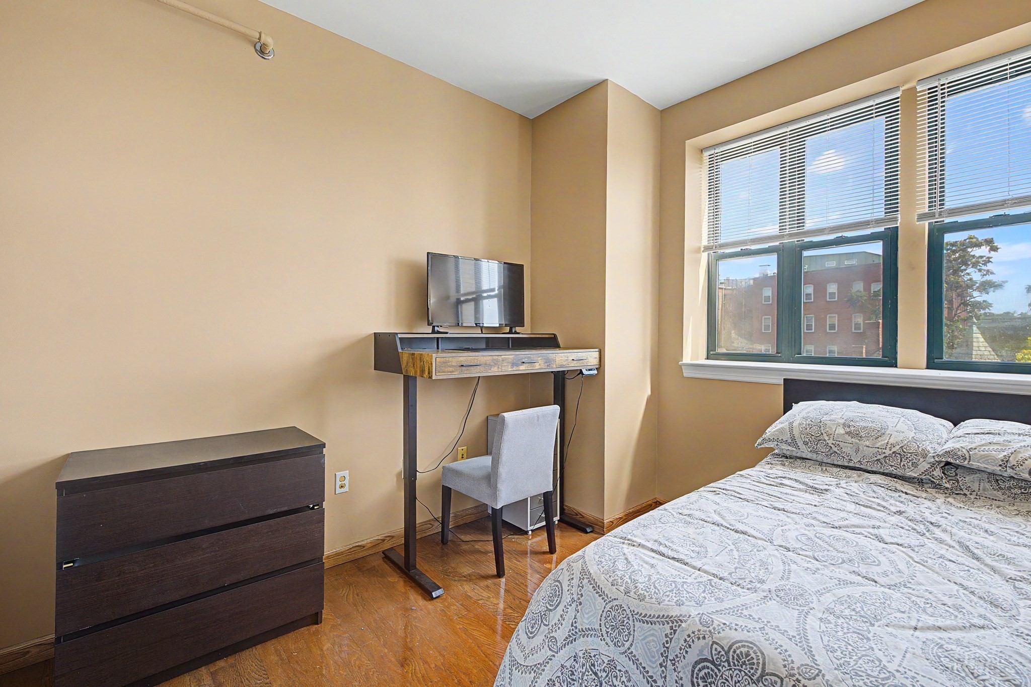 Bedroom with a patterned bedspread, desk and television, and large windows with blinds.