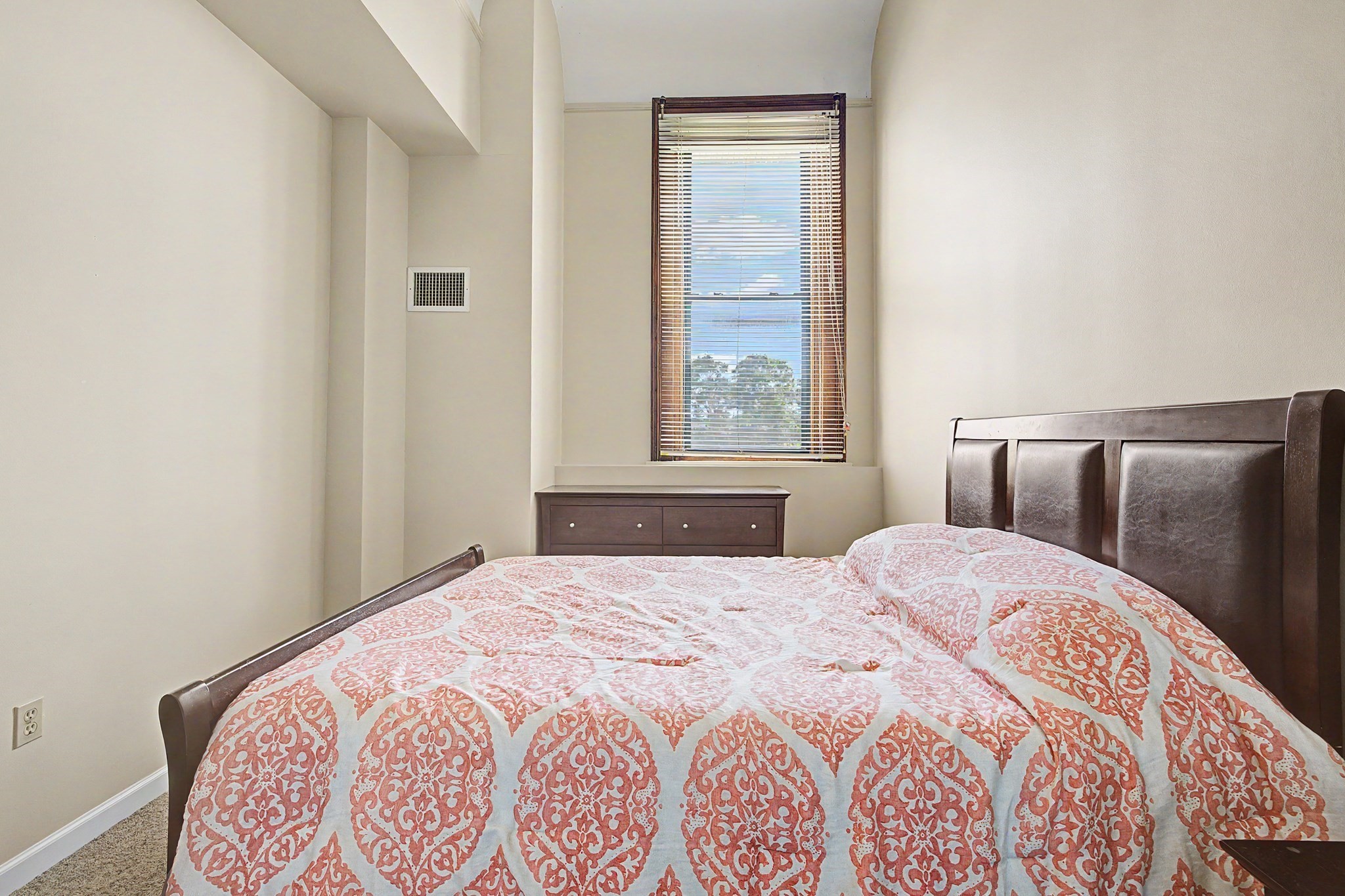 Bedroom with patterned bedding, a dark wood bed, and a tall window with blinds.