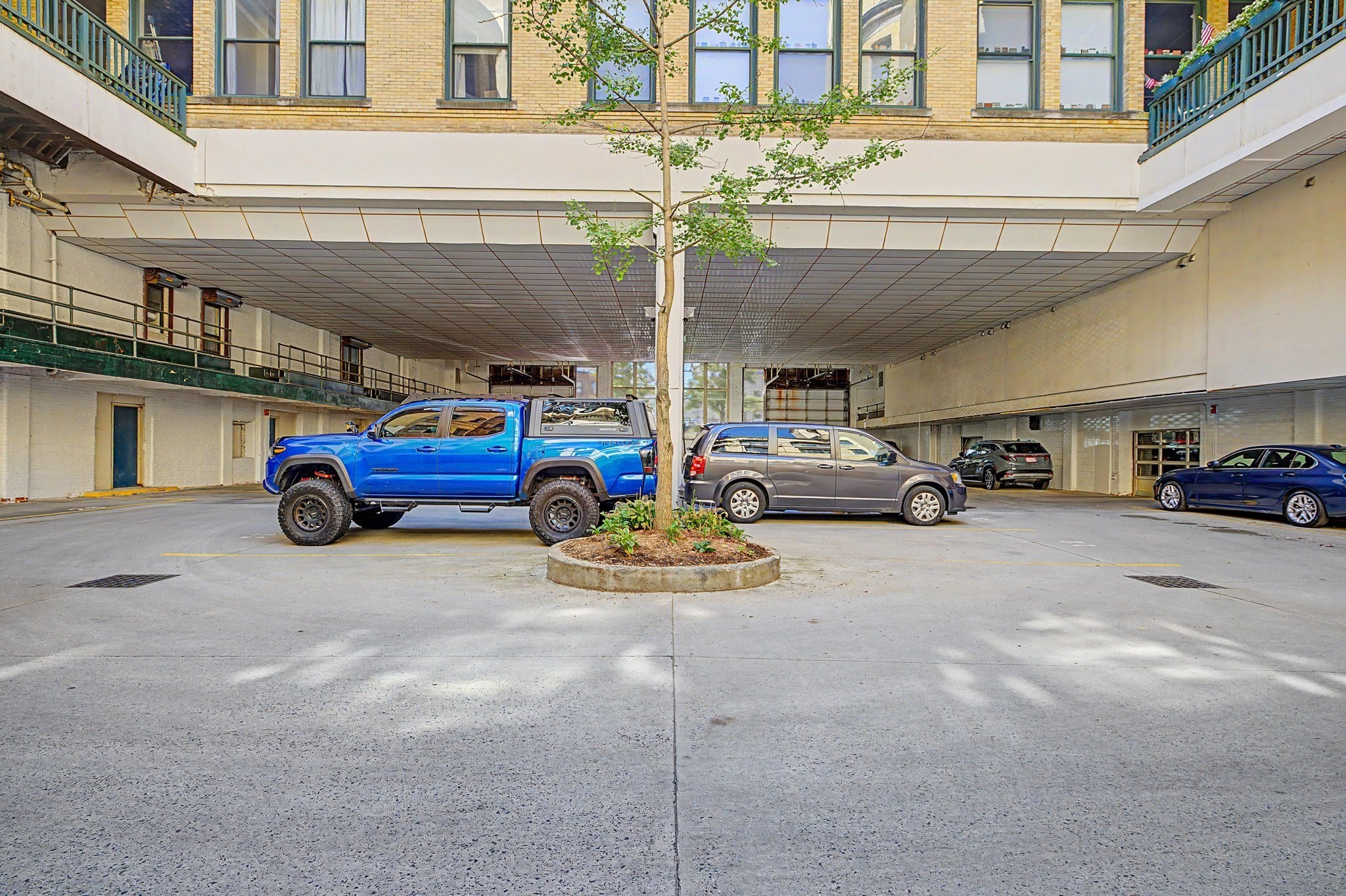 Covered parking area beneath a brick building, with parked cars and a tree in a circular planter.