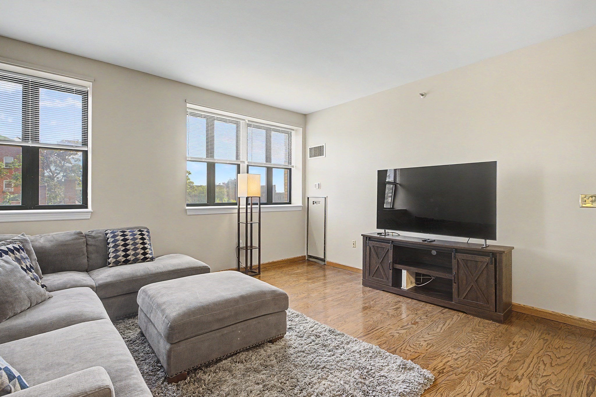 Living room with a gray sectional, large windows, hardwood floors, and a flat-screen TV.