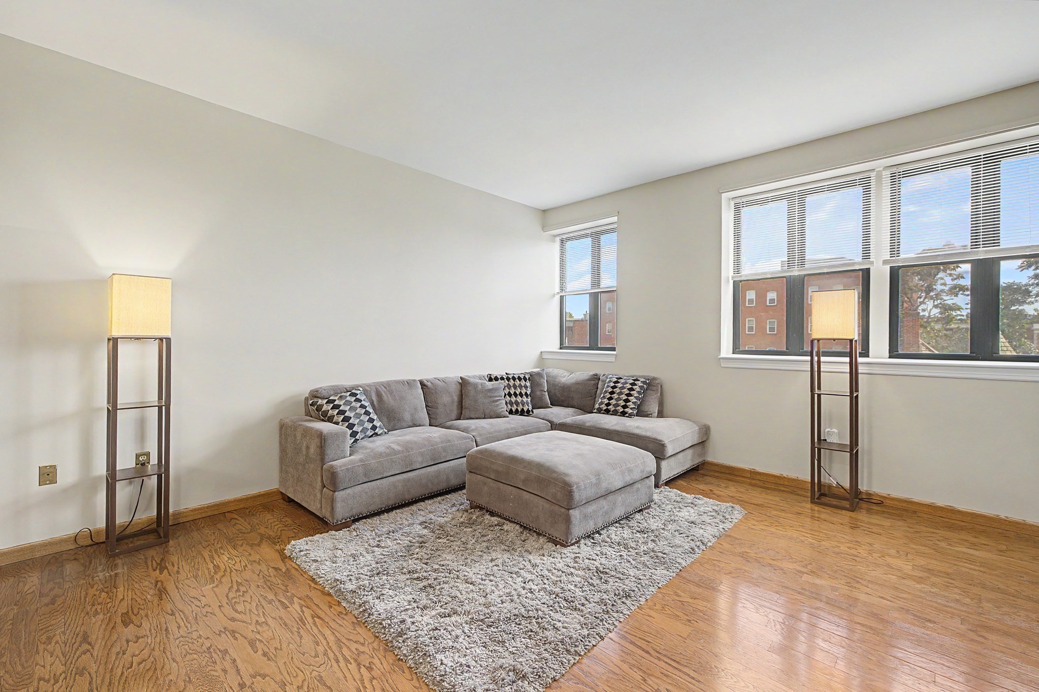 Living room with a gray sectional, ottoman, hardwood floors, and tall windows.