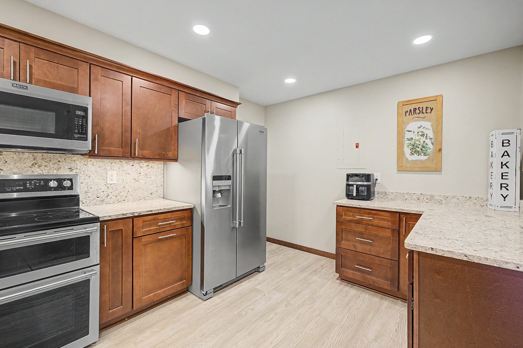 Kitchen with brown wood cabinets, stone counters, stainless appliances, and a bakery-themed sign.