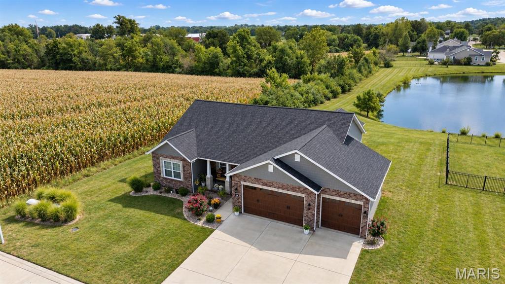 Aerial view of a brick home beside a pond and cornfield, with a two-car garage and landscaped lawn.