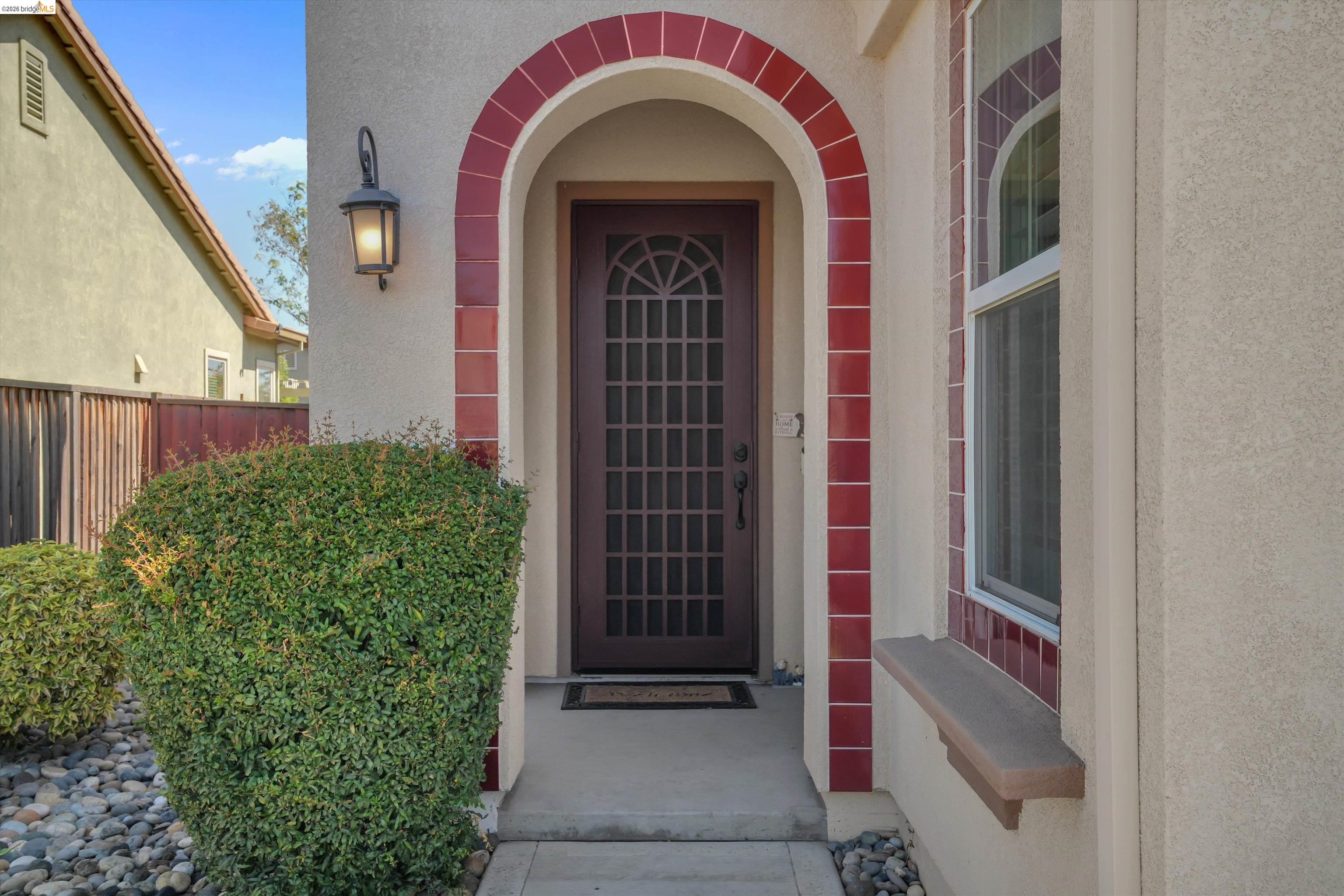 Arched front entry with a dark grid-paneled door, red tile trim, and trimmed shrubs.
