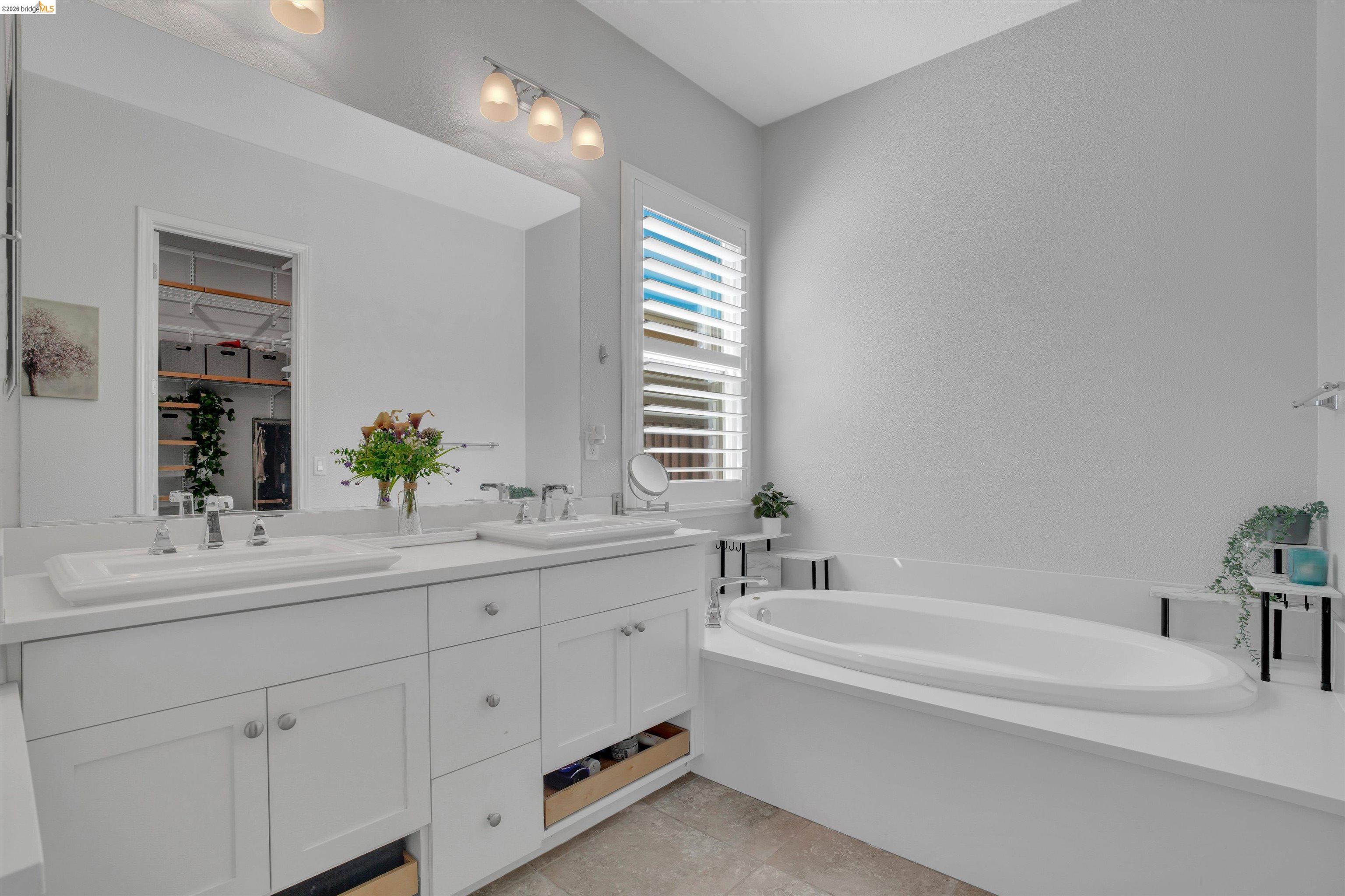 Bright bathroom with a double-sink vanity, shuttered window, and large corner bathtub.