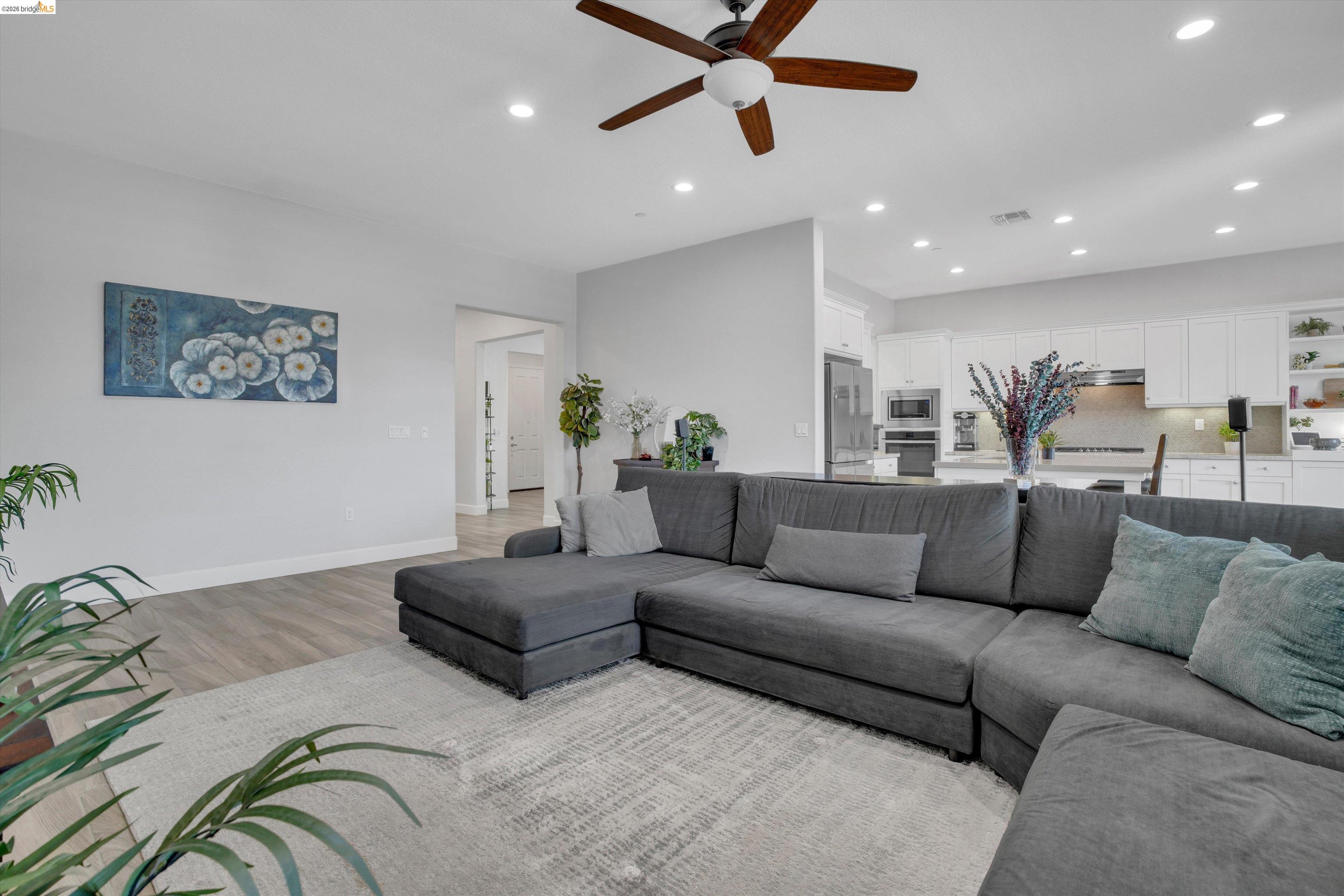 Open living room and kitchen with a gray sectional, white cabinetry, and a wood-blade ceiling fan.